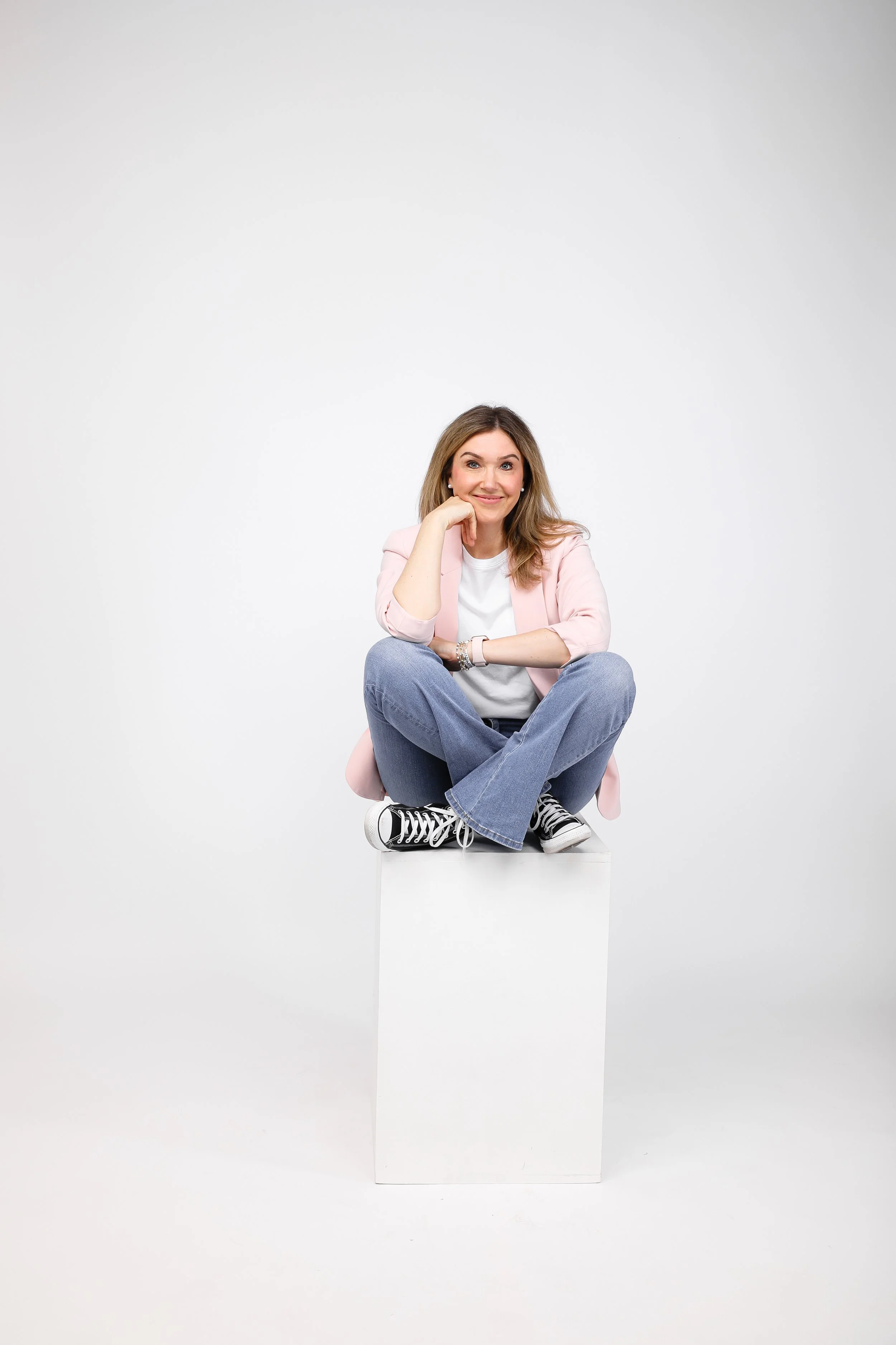 Author and CEO Jamie Moraga siting criss cross with pink blazer, jeans, and Chuck Taylor hightops, hand on chin