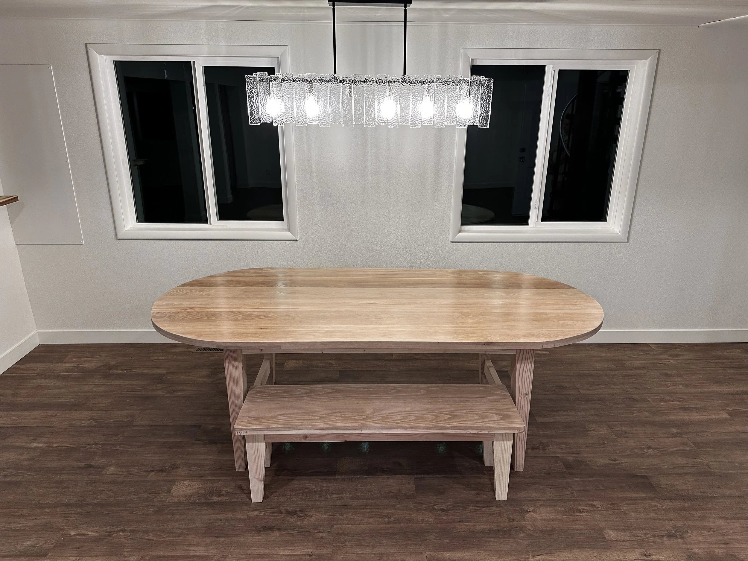 A wooden table with a shelf beneath, positioned in front of two windows on a light-colored wall, with a chandelier hanging above.