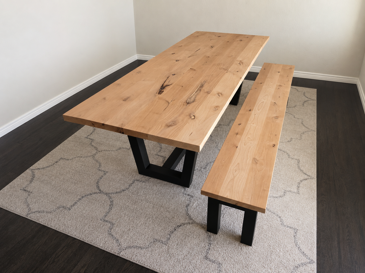A wooden table with a matching bench on a patterned rug in a room with light-colored walls and dark hardwood floors.