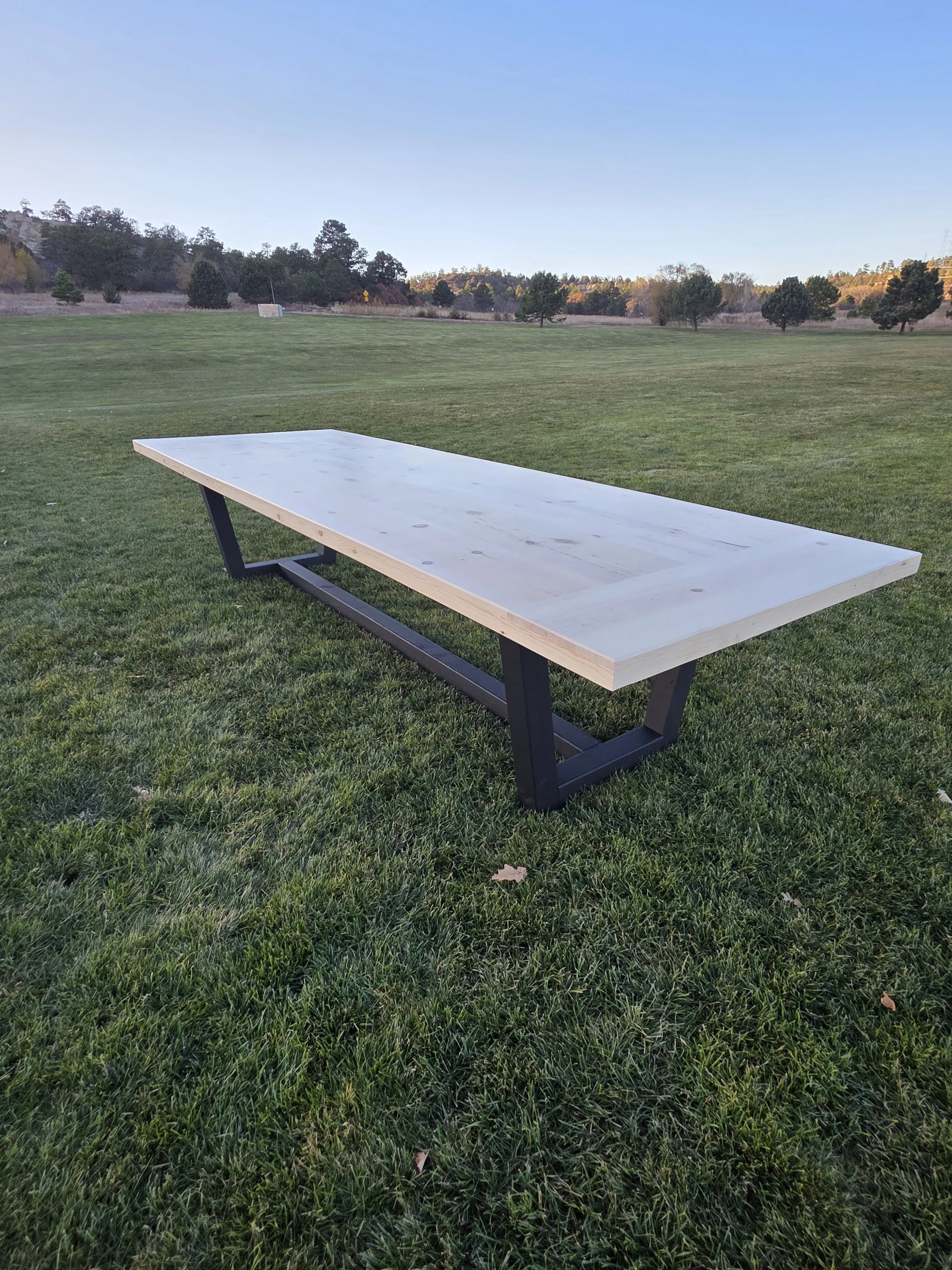 Empty wooden table with black metal legs on green grass in an open park with trees and clear blue sky in the background.