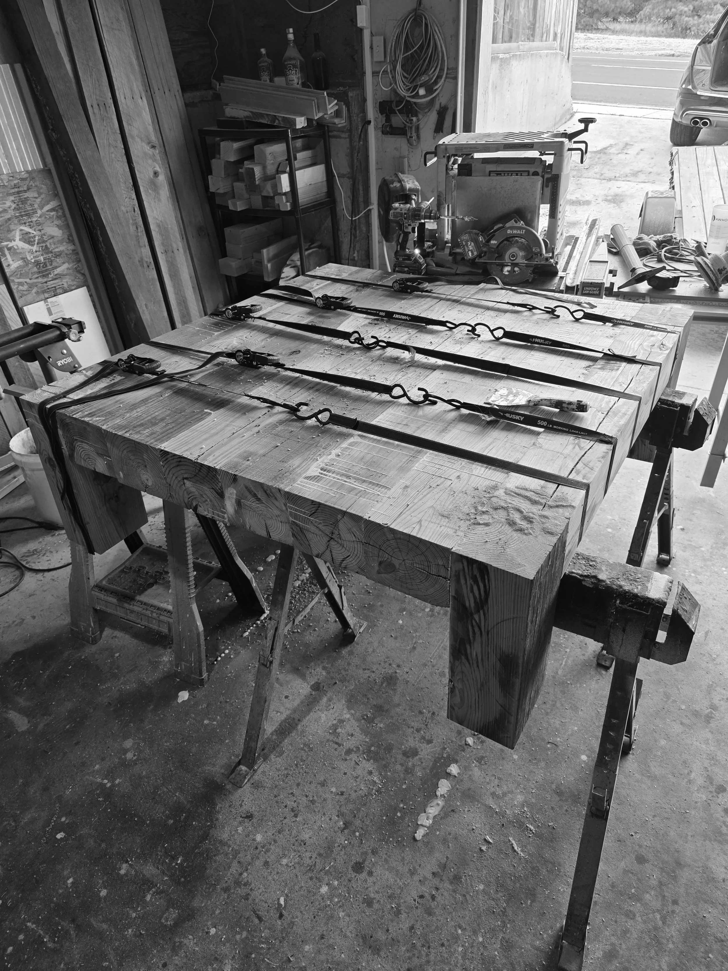 A cluttered woodworking workshop with a large wooden workbench, power tools, and various supplies. The workbench holds several straps, a drill, and a circular saw, with woodworking supplies on a shelf and hoses hanging on the wall. An open garage door reveals the outdoors and a parked vehicle.