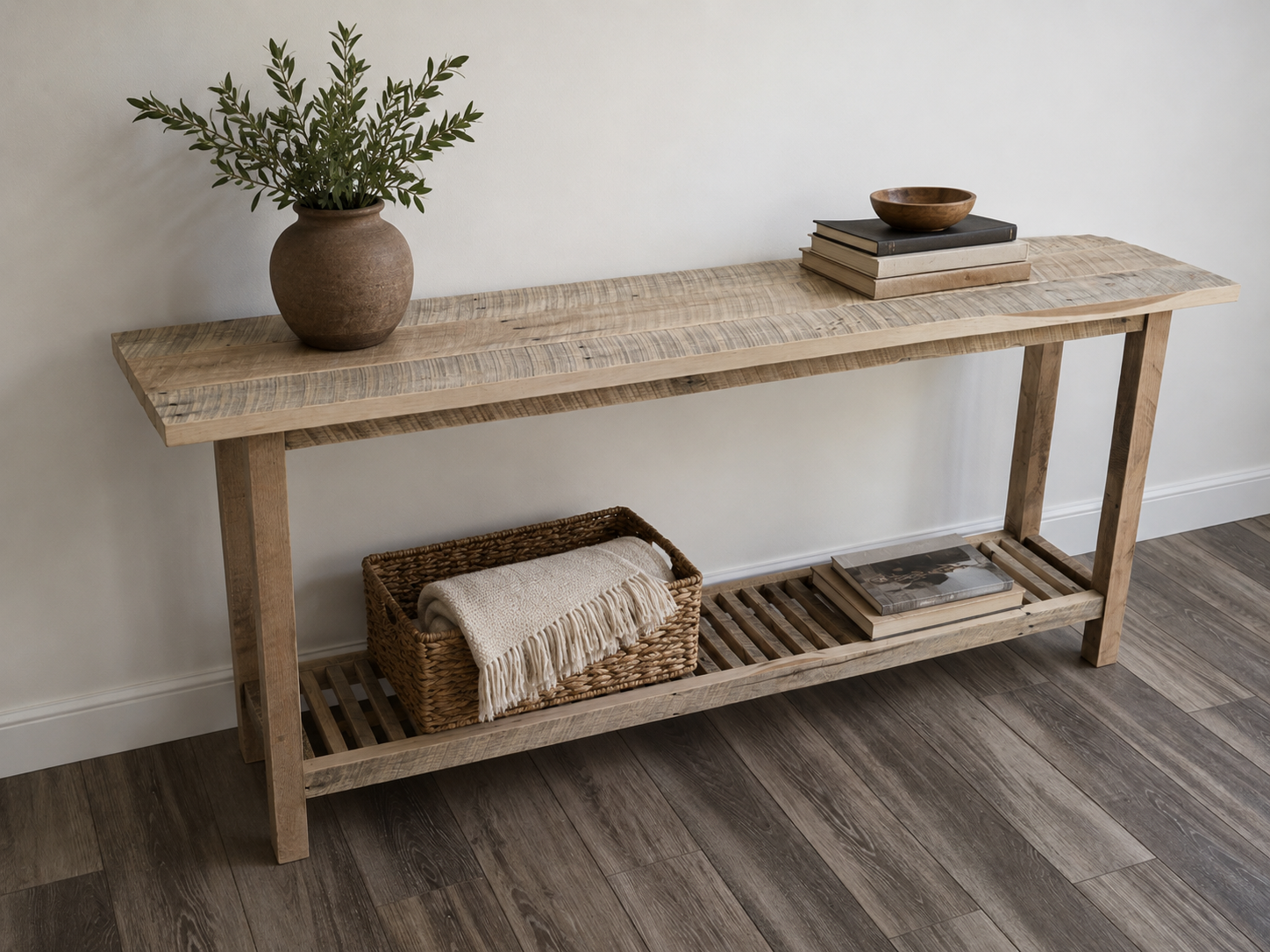 A light wooden console table with a slatted lower shelf, a ceramic vase with green foliage on the left, and a stack of books with a small wooden bowl on top on the right, against a plain white wall, with a dark wooden floor.