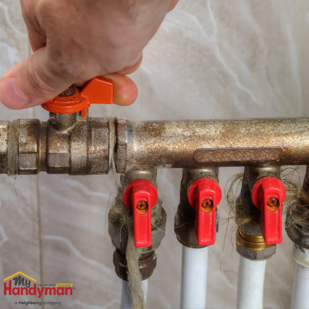 A hand turning an orange shutoff lever on a corroded brass water manifold with red valve handles, demonstrating main water shutoff before travel in Saline, Michigan.