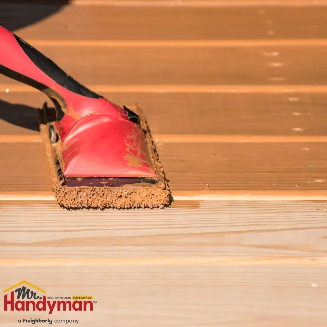 Close-up of a red foam pad applicator spreading a rich brown wood stain across freshly cleaned cedar deck boards in direct sunlight, representing the deck staining and sealing surface protection