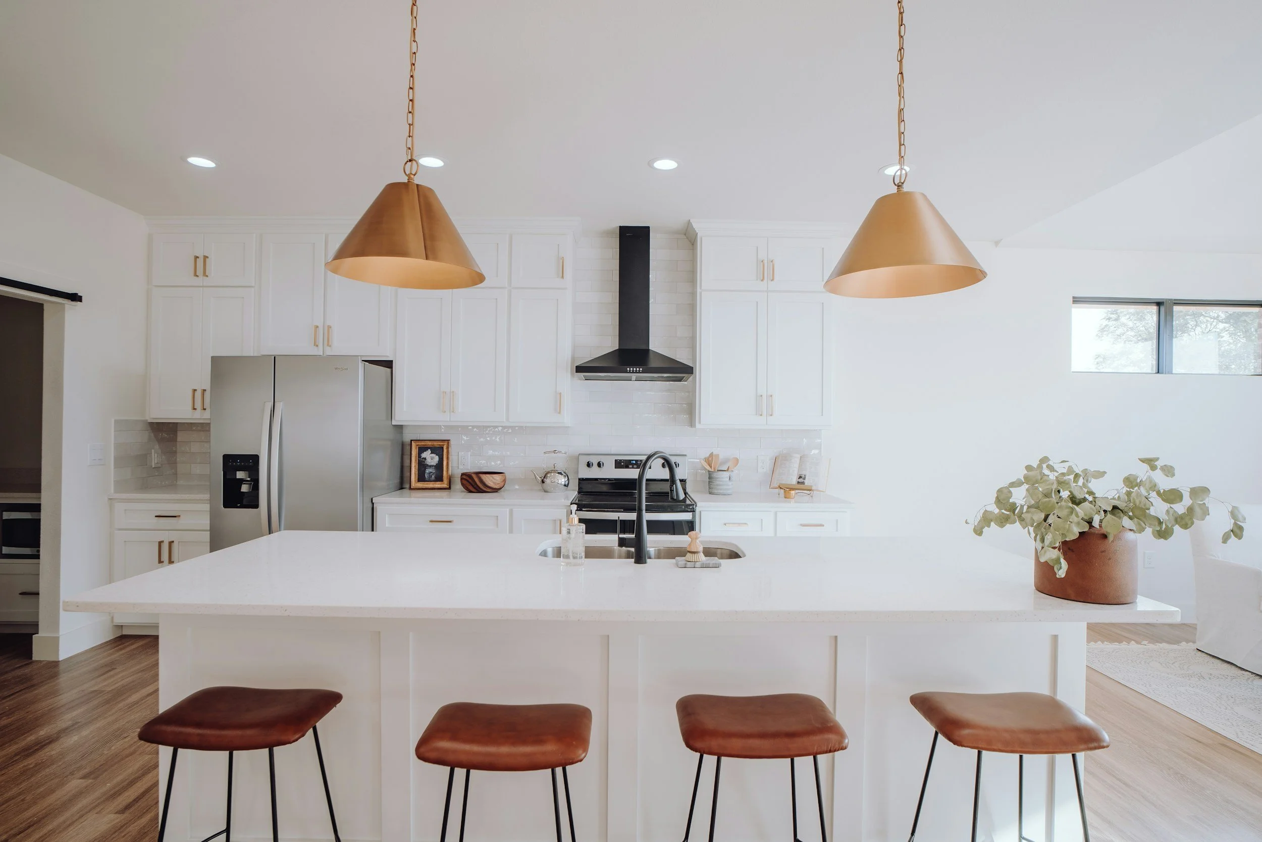 Modern kitchen with white cabinets, a stainless steel refrigerator, black range hood, and a white countertop island with a sink. Two copper pendant lights hang from the ceiling, and a potted plant on the island. There is a window on the right side, and wooden barstools with brown cushions at the island.