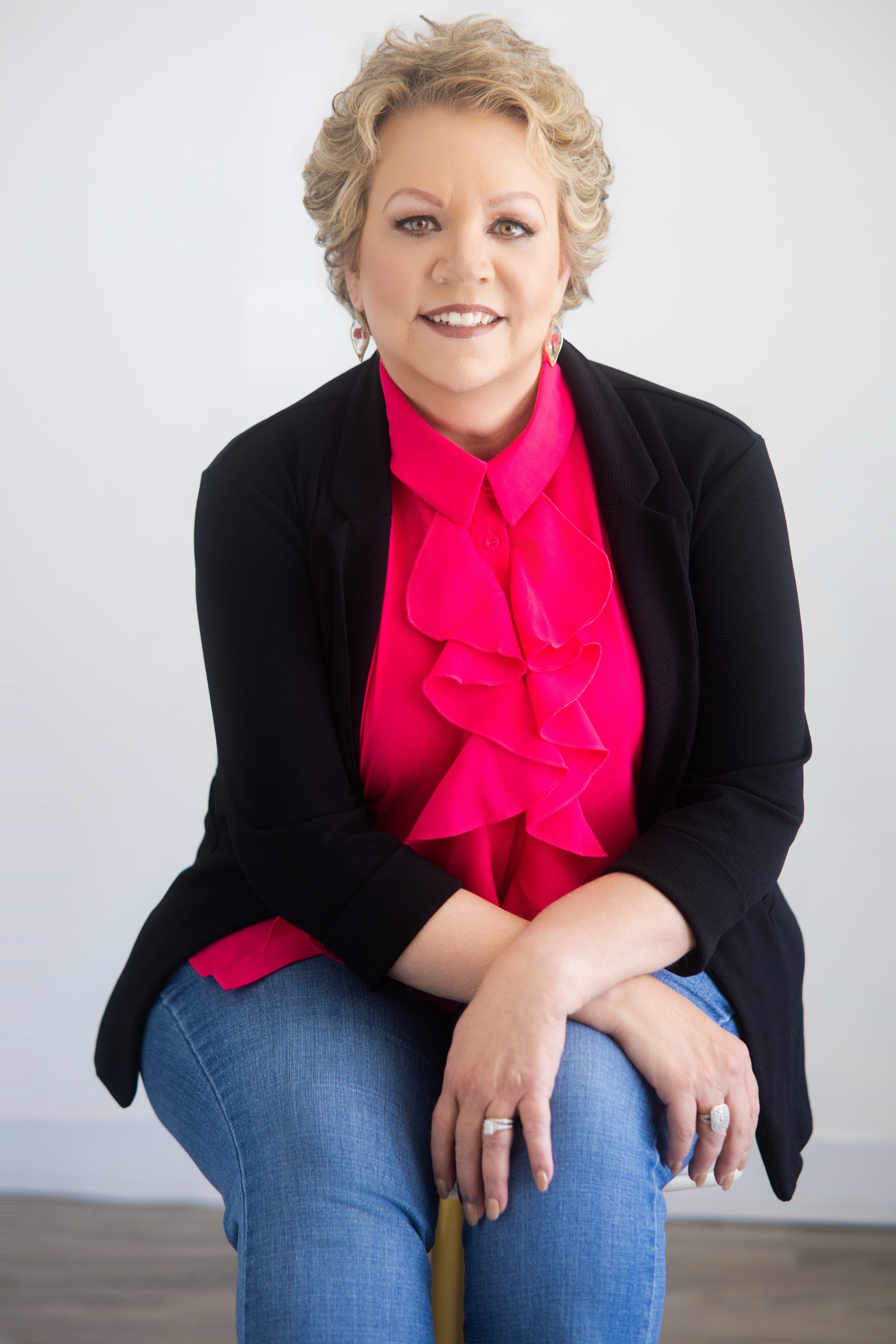Melody Hackett with short, curly blonde hair, wearing a bright pink blouse, a black jacket, and blue jeans, sitting on a chair with her arms crossed and hands resting on her lap in front of a plain light gray background. Richmond VA central based