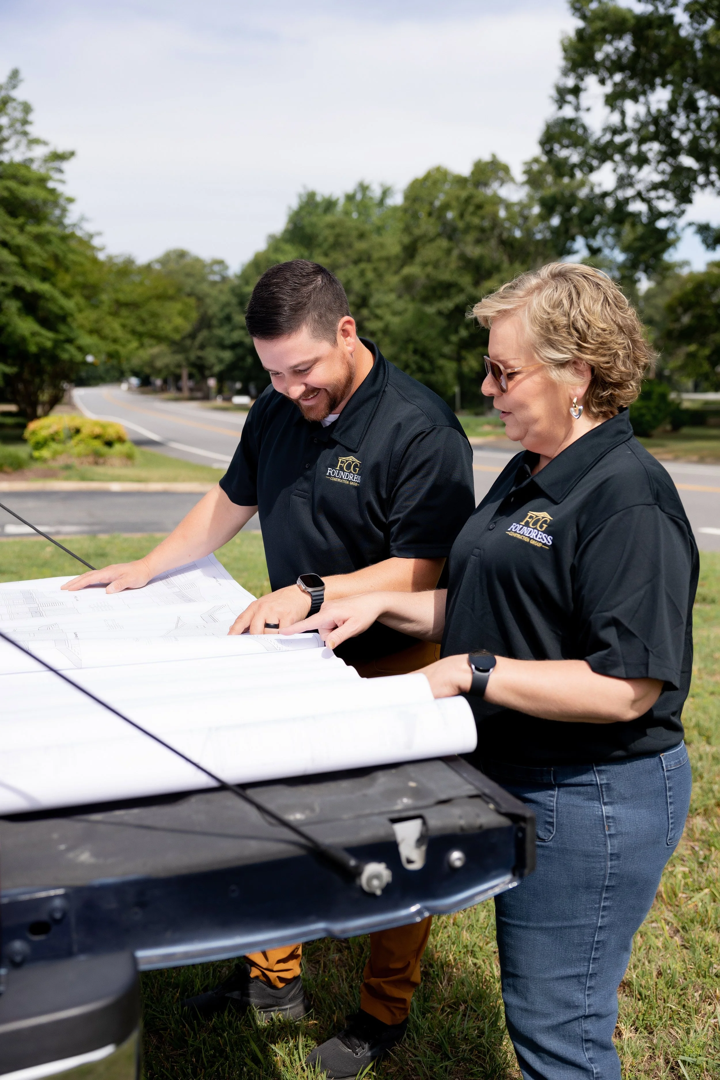 Two people wearing black shirts with 'FG Founders' logo reviewing blueprints outdoors on a grassy area with trees and cloudy sky in the background. Richmond VA central based home construction company, specializing in new homes, renovations, additions