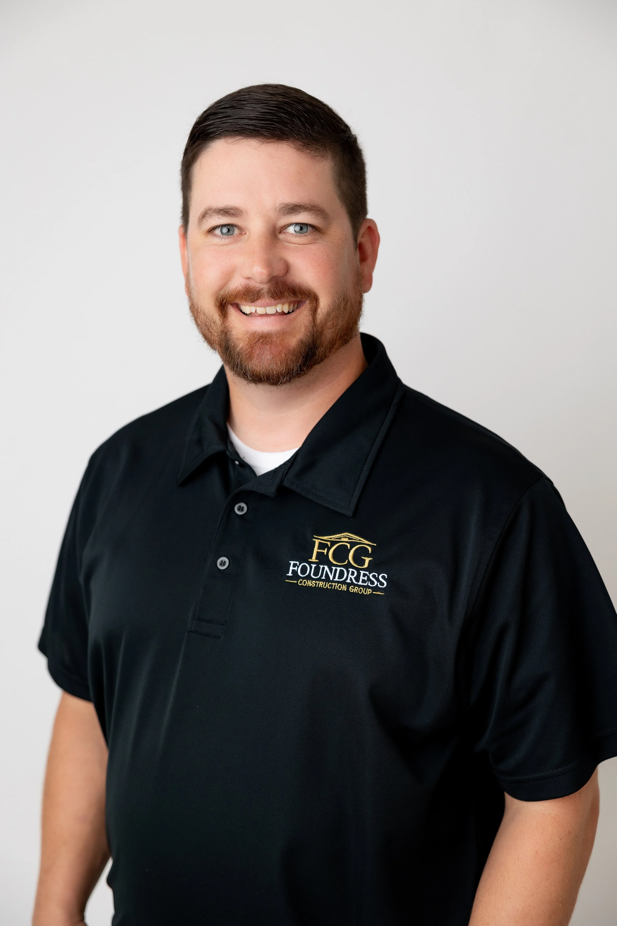 Blake Henry with short dark hair, blue eyes, and a beard, smiling and wearing a black polo shirt with a logo that reads 'FCG FOUNDRESS CONSTRUCTION GROUP'. Richmond VA central based home construction company, specializing in new homes, renovations, a