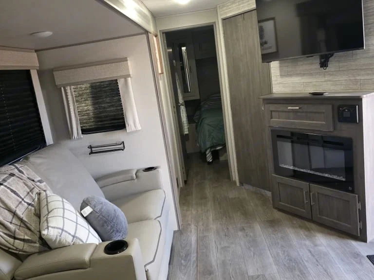 Interior of a compact living space with a cream-colored couch, pillows, a small window with curtains, a door leading to a bedroom, a wall-mounted TV, and a built-in electric fireplace.