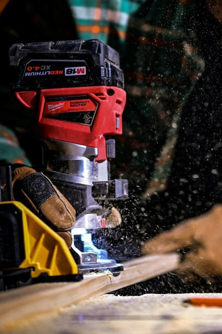 A person operating a red Milwaukee router tool with a black battery attached, working on a wooden surface with wood dust flying around.