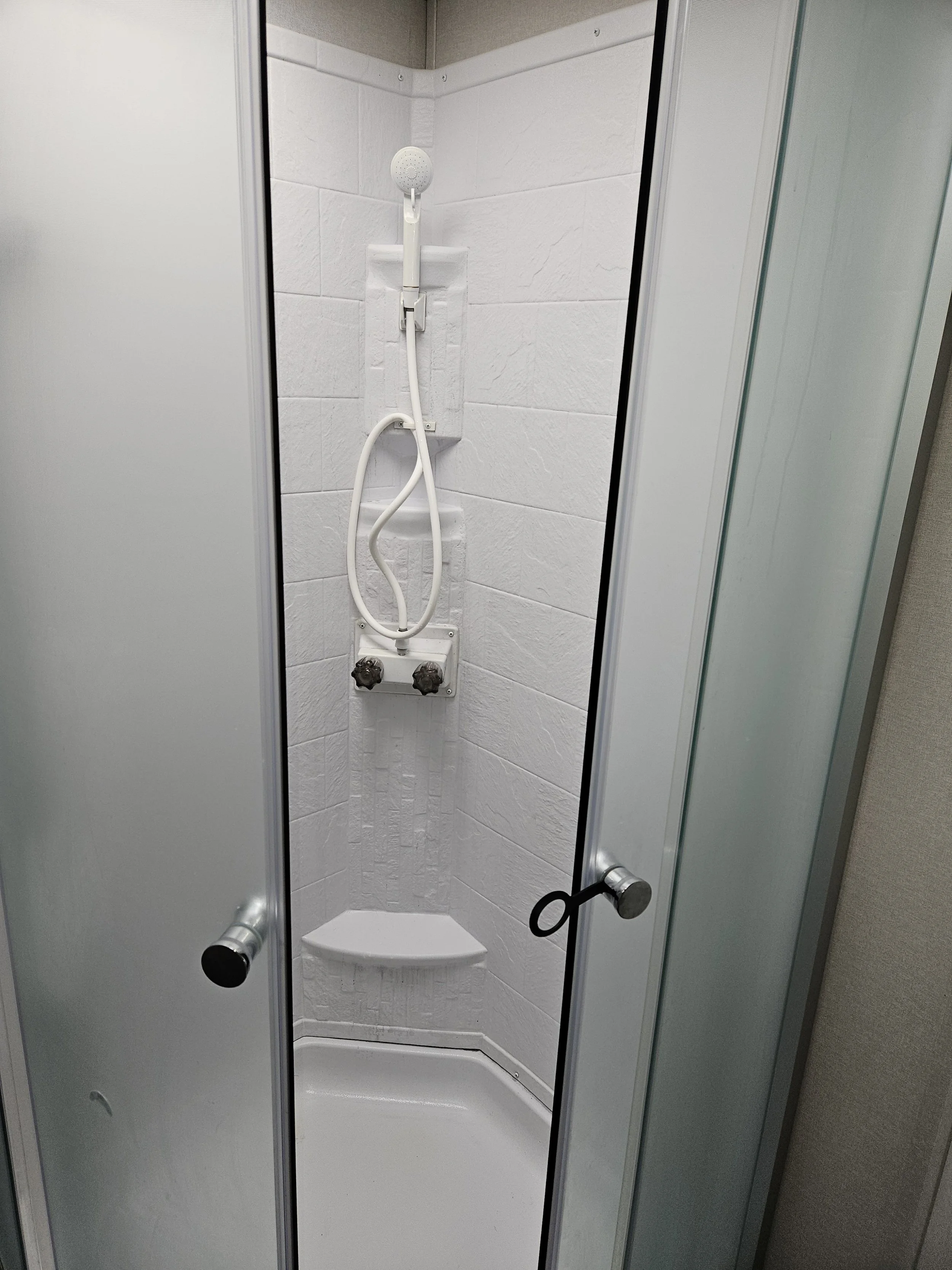 Inside a small shower stall with white textured walls, a showerhead, and two temperature control knobs. The stall has a corner shower seat and a glass door with metal handles.