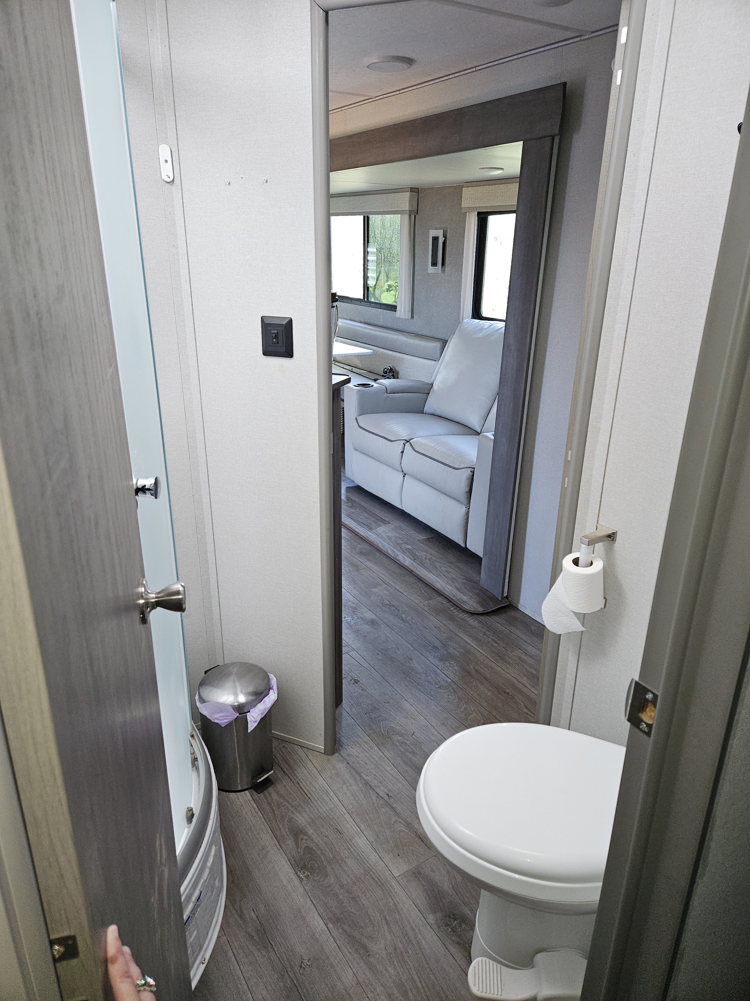 View inside a small bathroom with a washing machine on the left, a trash bin with a purple bag, a toilet in the foreground, a white sofa near window, and various fixtures and windows in a camper or RV interior.