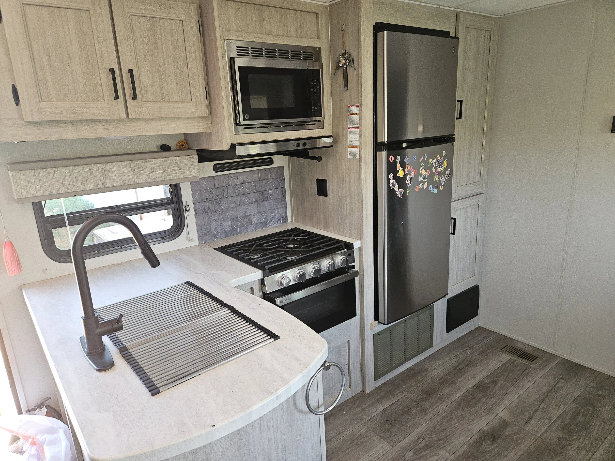 Kitchen inside a camper or small home, showing a window with a small sink, a stove with an oven, a microwave, and a refrigerator decorated with colorful magnet letters, with light-colored cabinetry and wood-look flooring.