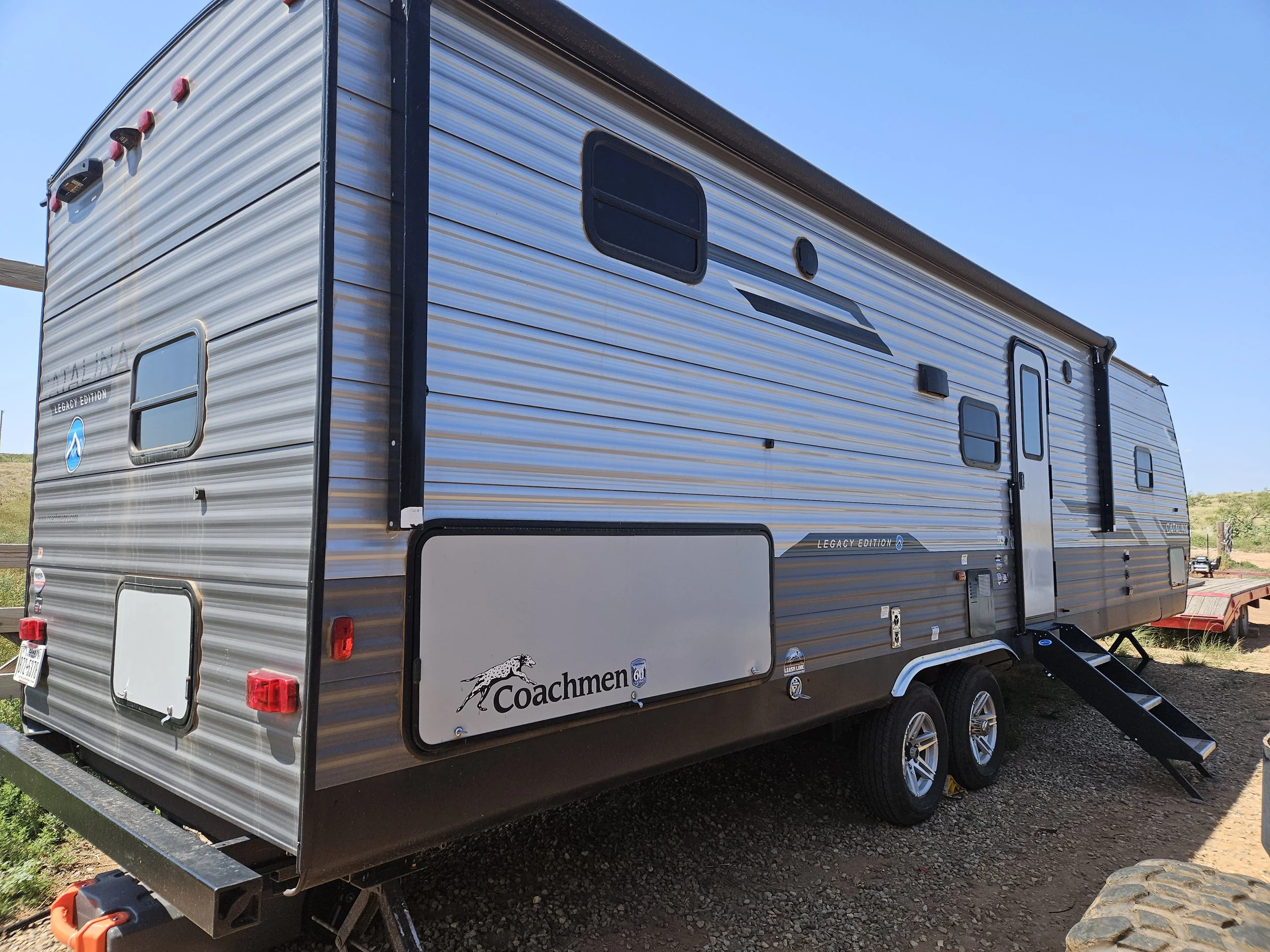 A modern silver travel trailer with black accents and multiple windows, parked on a gravel lot under a clear blue sky. The trailer has a Coachmen logo with a running deer and the words "Coachmen" on the side.