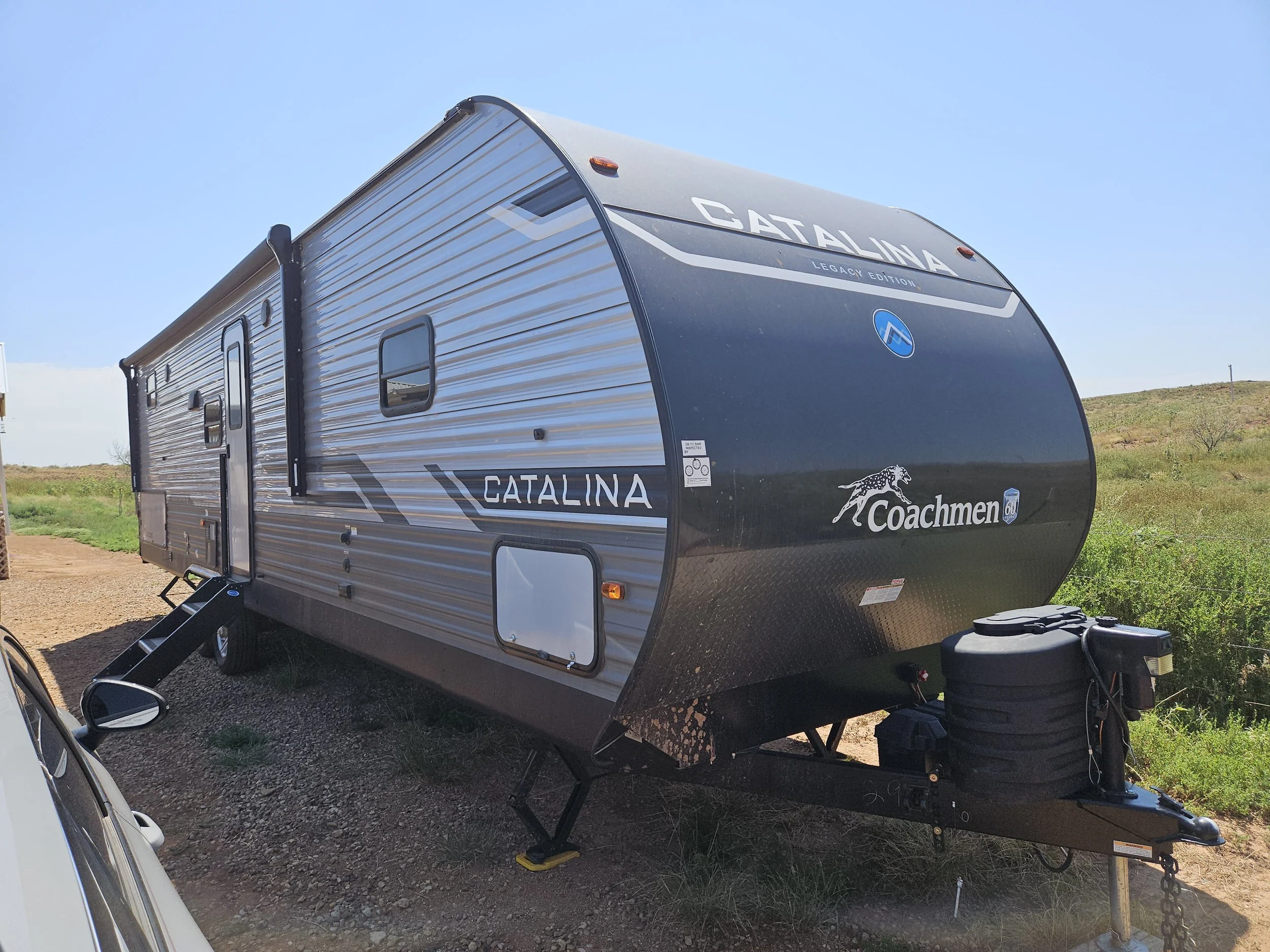 A grey and black Coachmen Catalina travel trailer with branding and logo, parked on a gravel lot with green fields and a blue sky in the background.