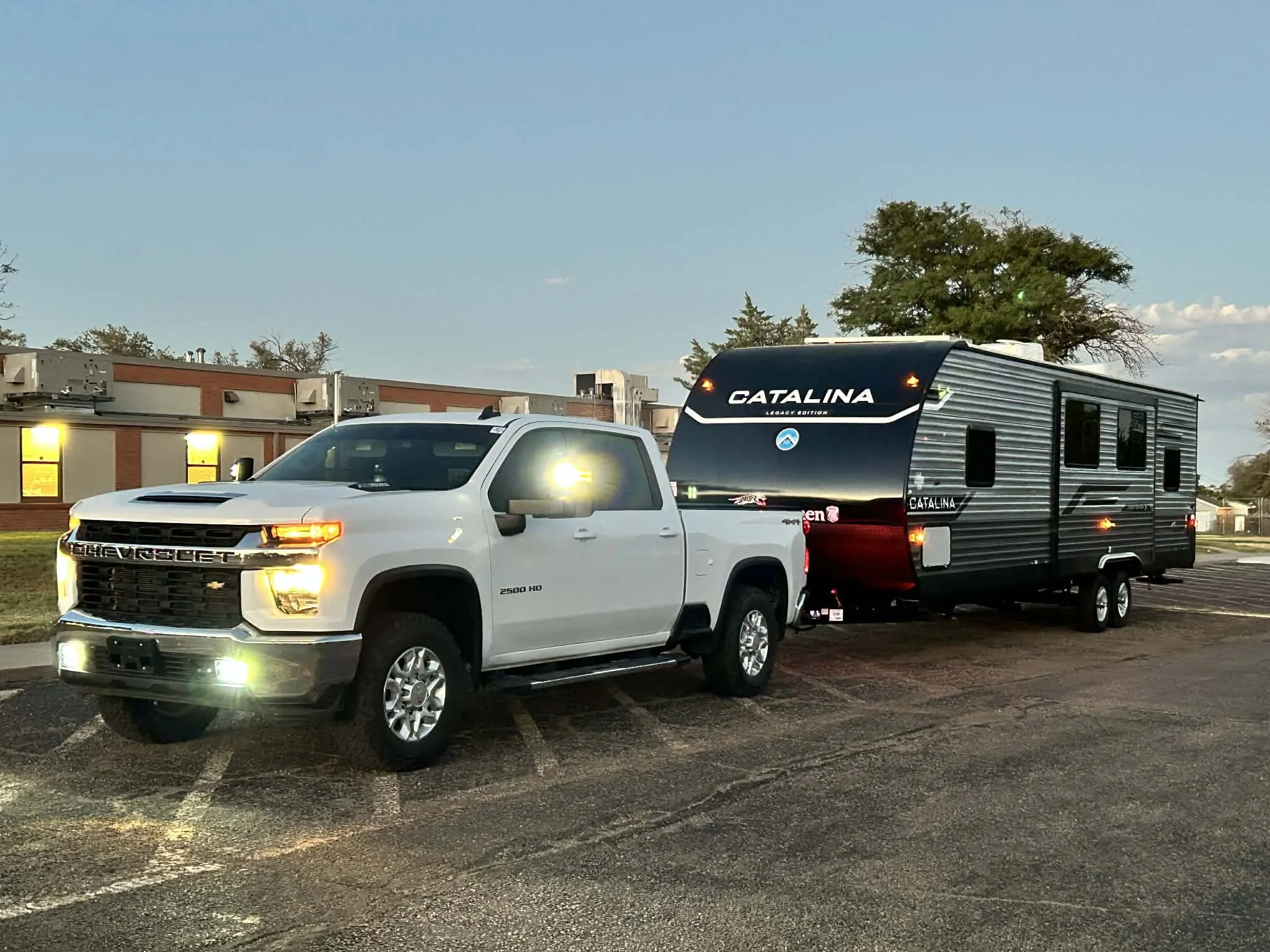 A white Chevrolet pickup truck pulling a black Catalina travel trailer parked in a parking lot with a building and trees in the background, during early evening.
