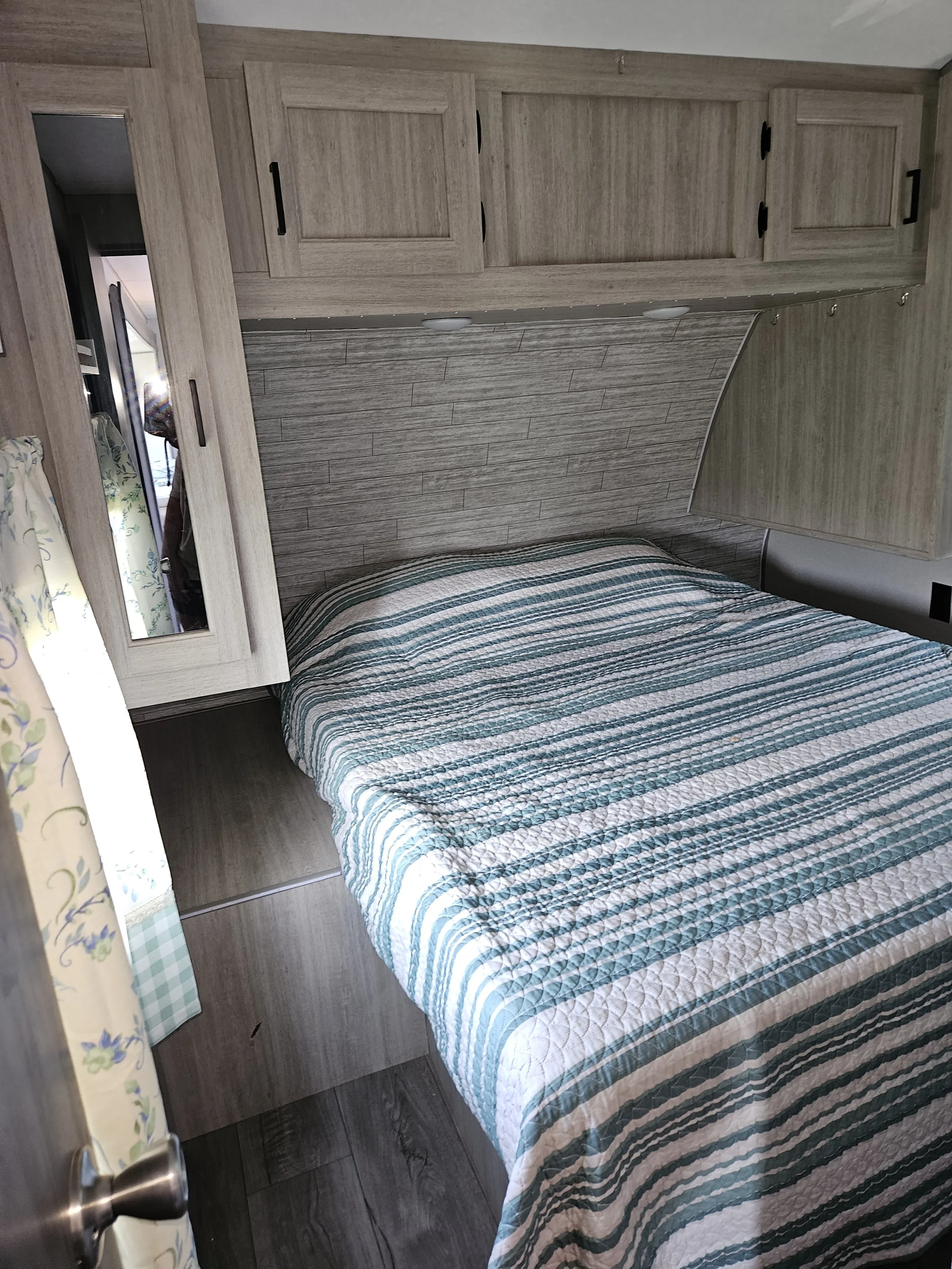 Interior of a bedroom in a recreational vehicle, showing a bed with a striped quilt, wooden cabinetry overhead, a mirror on the left, and a floral curtain partially covering a window.