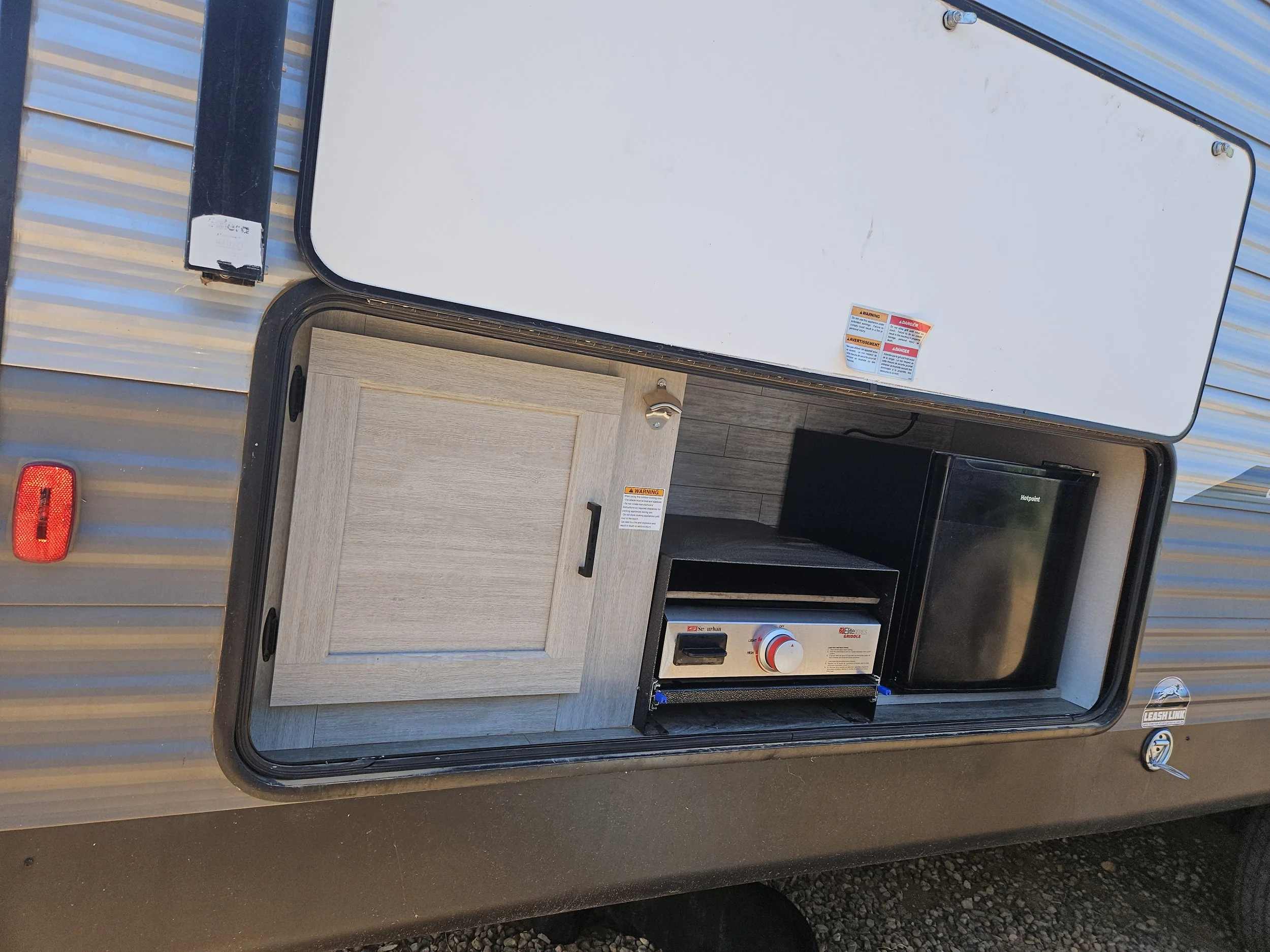 A silver travel trailer with an open side door revealing a small outdoor kitchen area with a grill, a wood panel drawer, and several warning stickers on the white exterior wall.