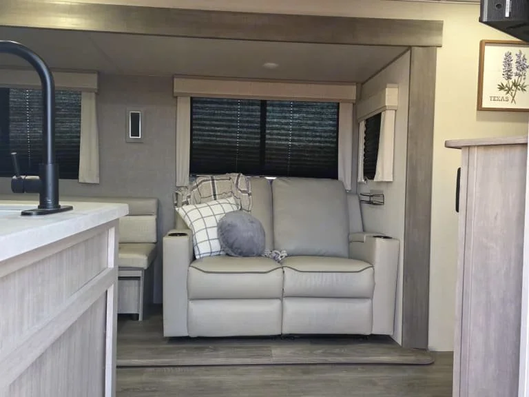 Interior view of a cozy living area with a beige sofa, plaid pillows, a window with black blinds, and a wooden cabinet in a neutral-toned room.