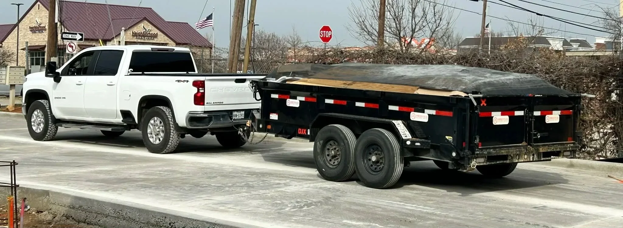 A white Chevrolet pickup truck towing a black trailer with a cover. The truck is parked on a paved road with a stop sign and buildings in the background.