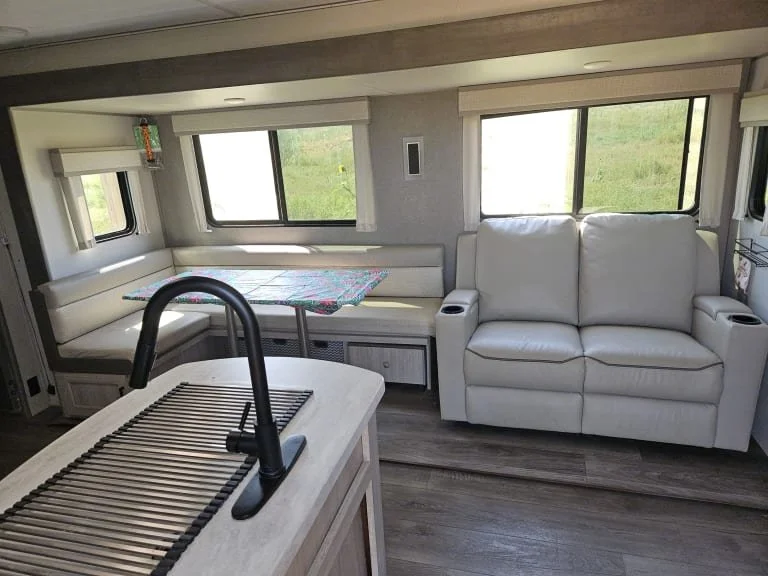 Interior of a camper with a white leather sofa, a dining area with a colorful tablecloth and built-in seating, windows showing green grass outside, and a kitchen island with a black faucet.