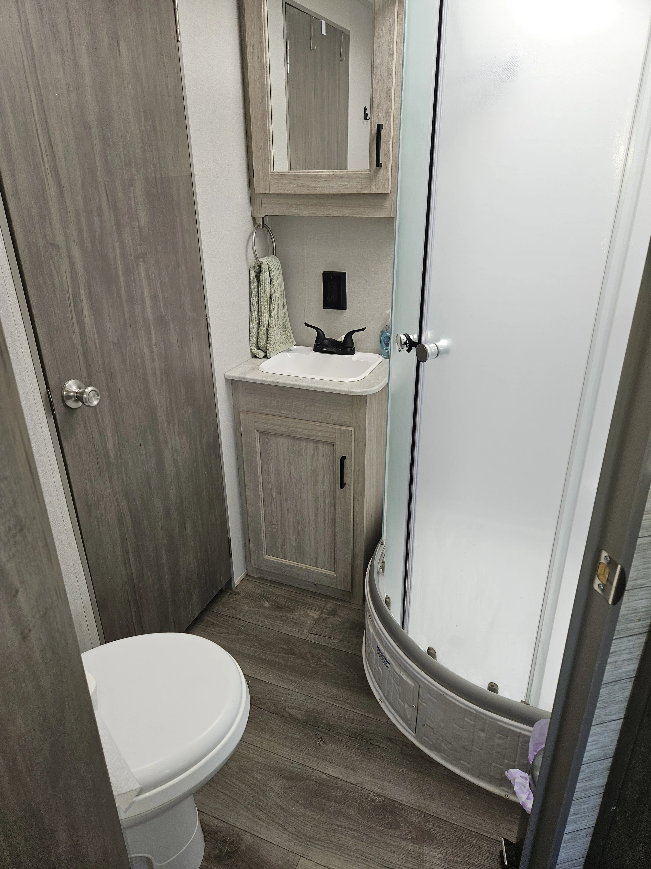 A compact bathroom with a toilet, a small sink with a black faucet, a mirror cabinet above the sink, a hand towel hanging on a ring, a black outlet on the wall, wooden cabinetry, and a curved shower enclosure with a frosted glass door on a wooden flo