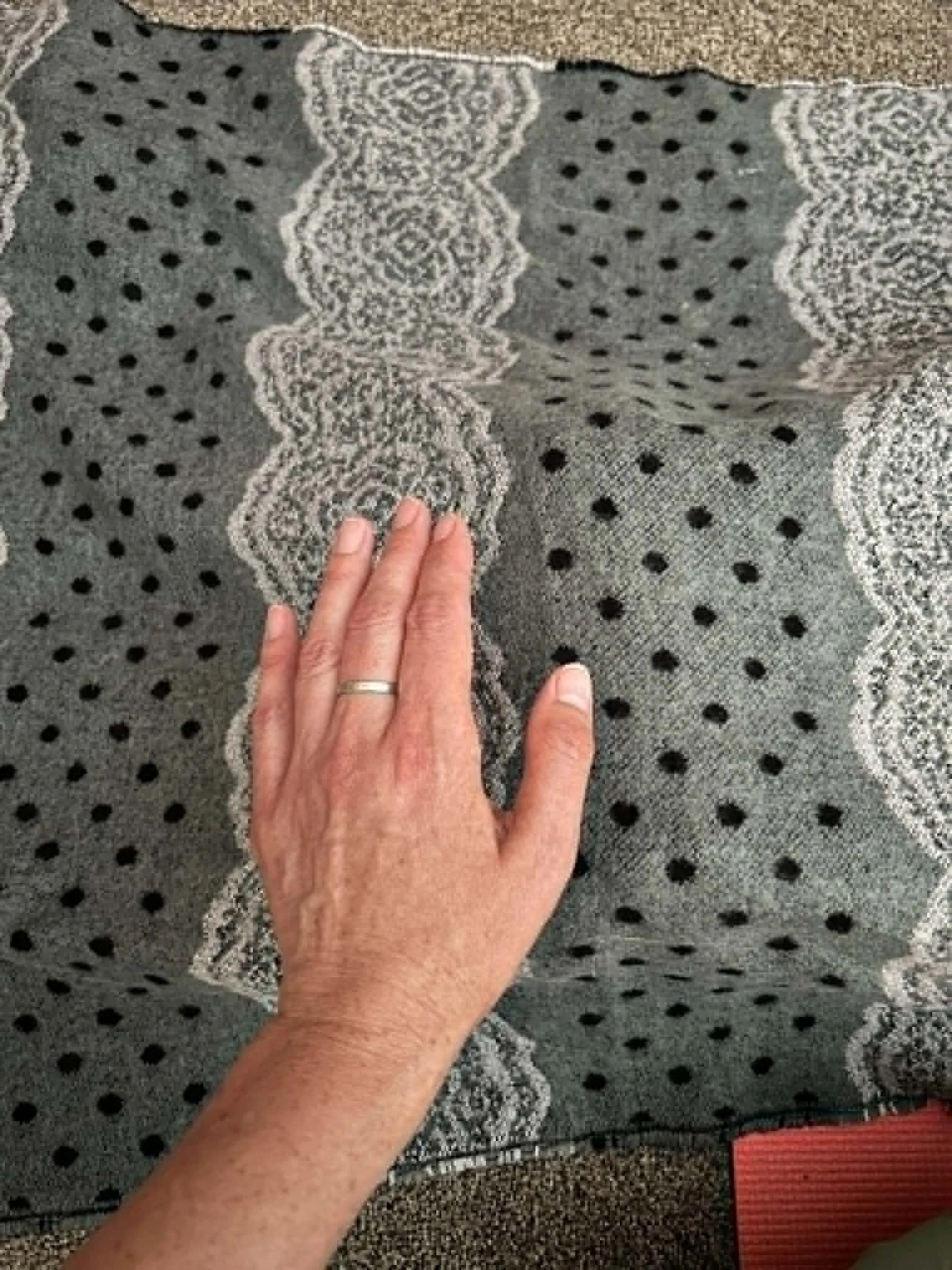 A person's hand is touching a patterned fabric with a wavy design and black polka dots. The fabric appears to be a carpet or rug.