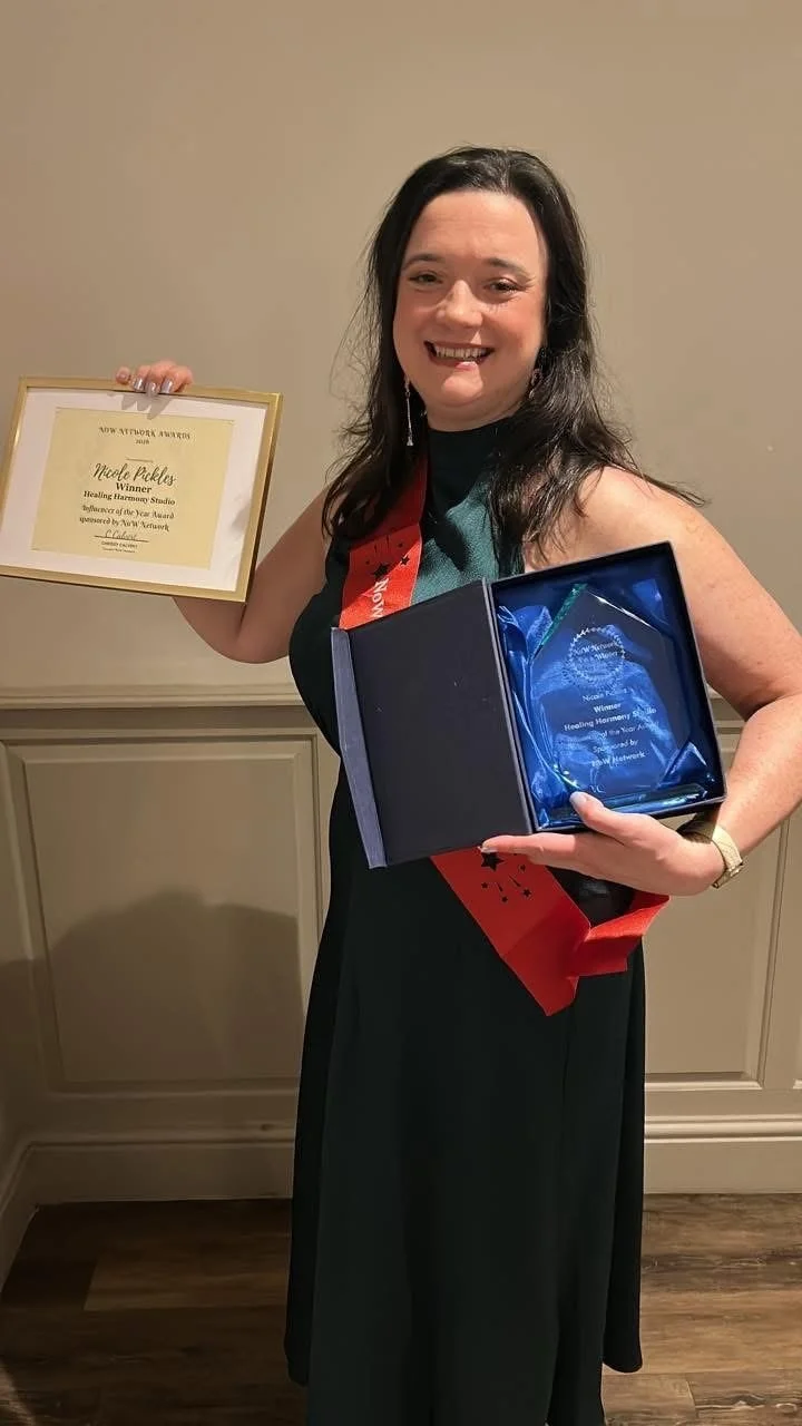 Woman holding a framed award certificate and a blue glass trophy, wearing a sash, at an award ceremony.
