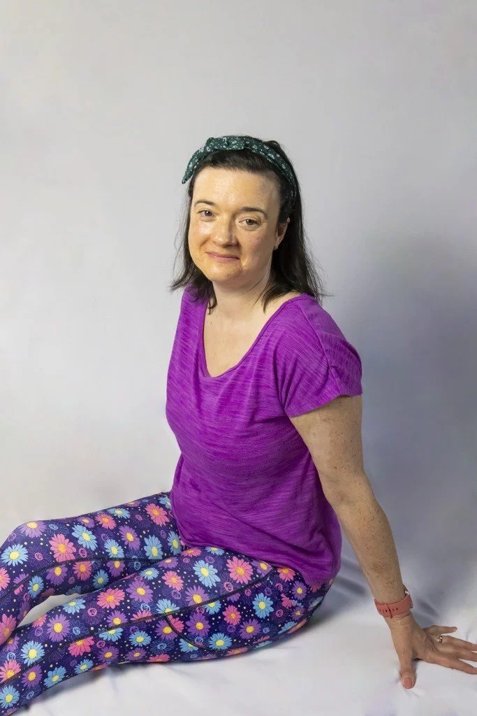 A woman with dark hair, wearing a purple top and colorful floral leggings, sitting on the floor against a plain background.