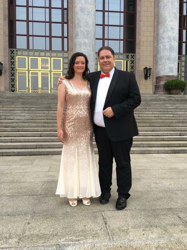 A man and woman dressed in formal attire standing on the steps in front of a large building with tall columns and glass doors, smiling at the camera.