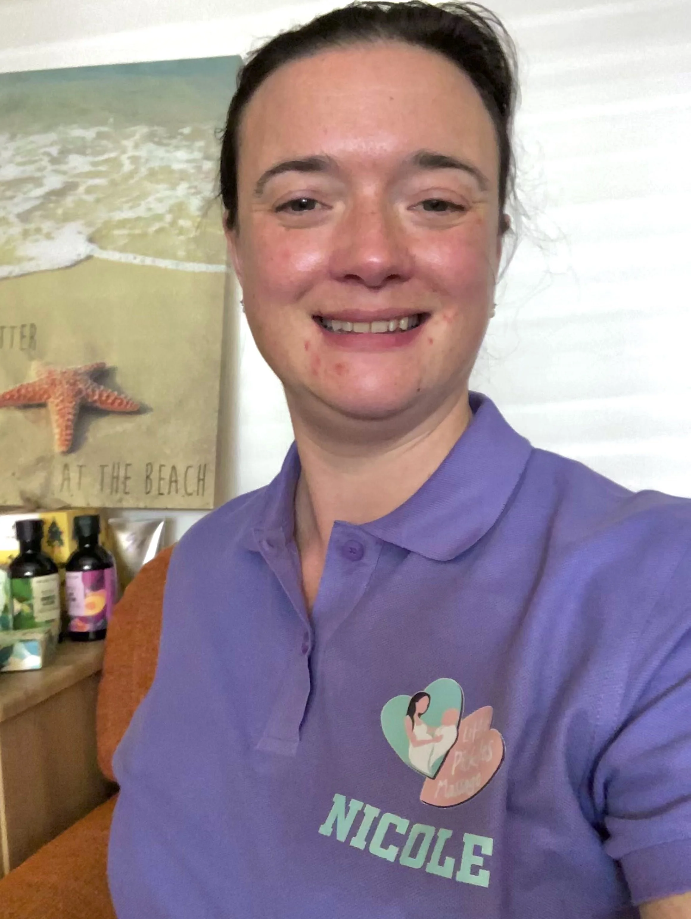 Close-up of a woman smiling, wearing a purple shirt with a graphic of a mother and child, behind a wooden counter with bottles and a poster with a starfish and beach scene.