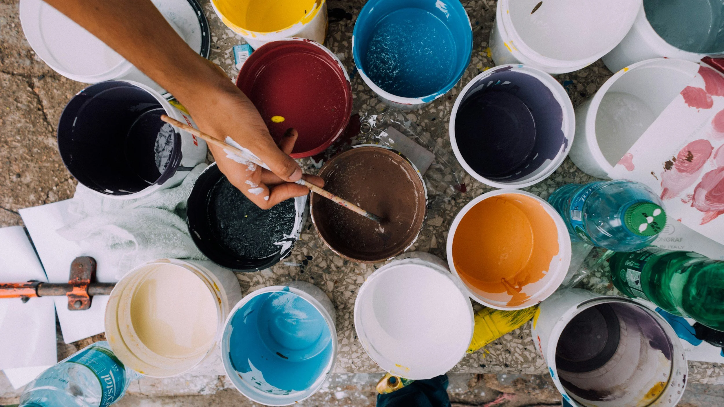 A person's arm using a paintbrush to mix brown paint in a bucket, surrounded by multiple open paint buckets in various colors, placed on a concrete surface.