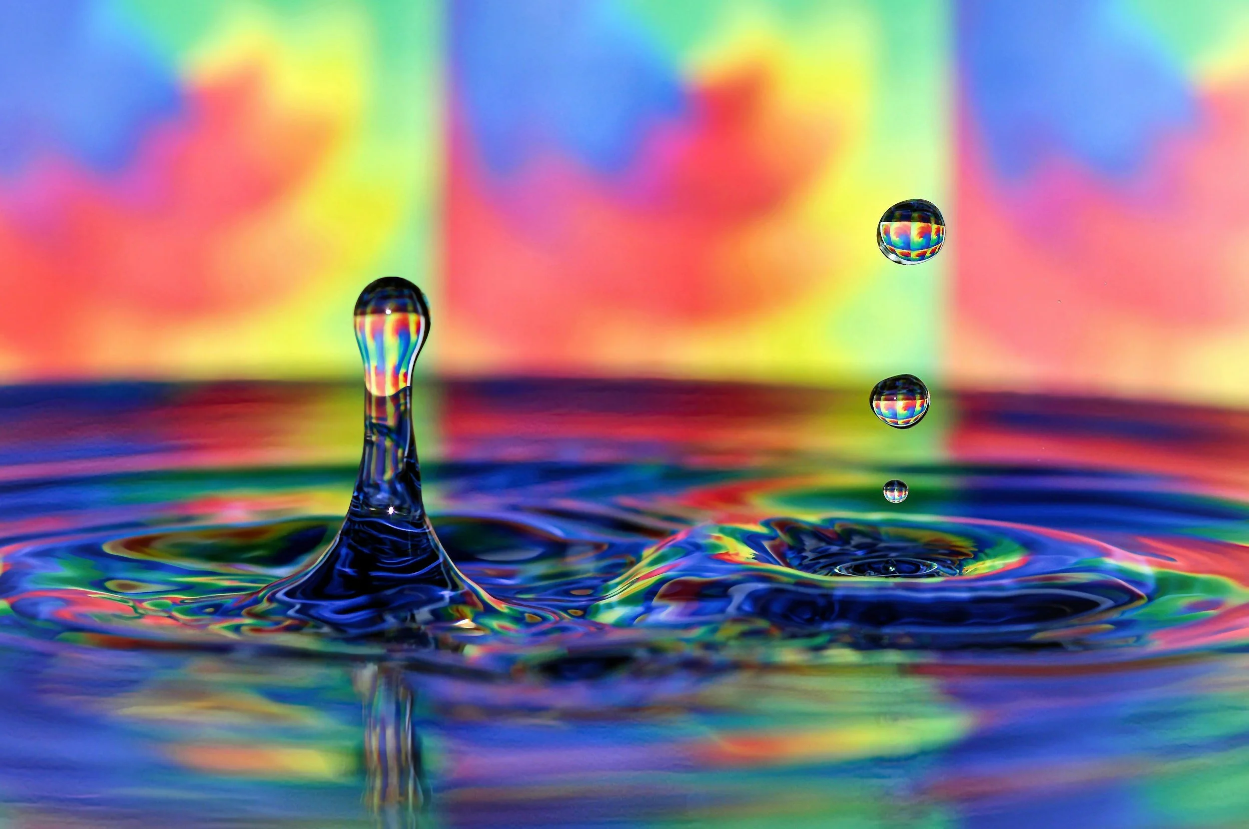 Close-up of water droplets falling into a colorful, iridescent surface, creating ripples and a splash, with a rainbow-like background.