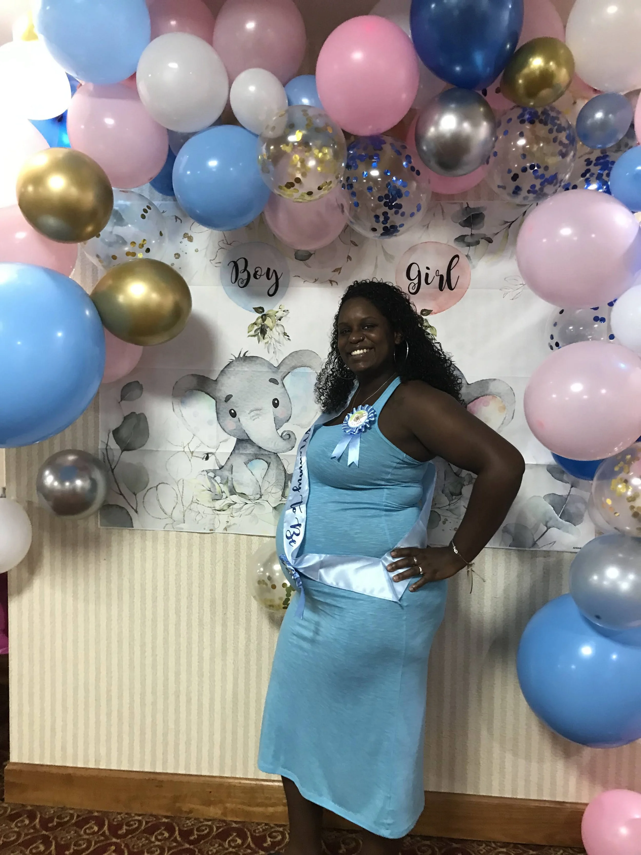 Woman smiling at baby shower, wearing blue dress and sash, surrounded by pink, blue, white, gold, and confetti-filled balloons, with a backdrop that says 'Boy' and 'Girl' and features an elephant illustration.