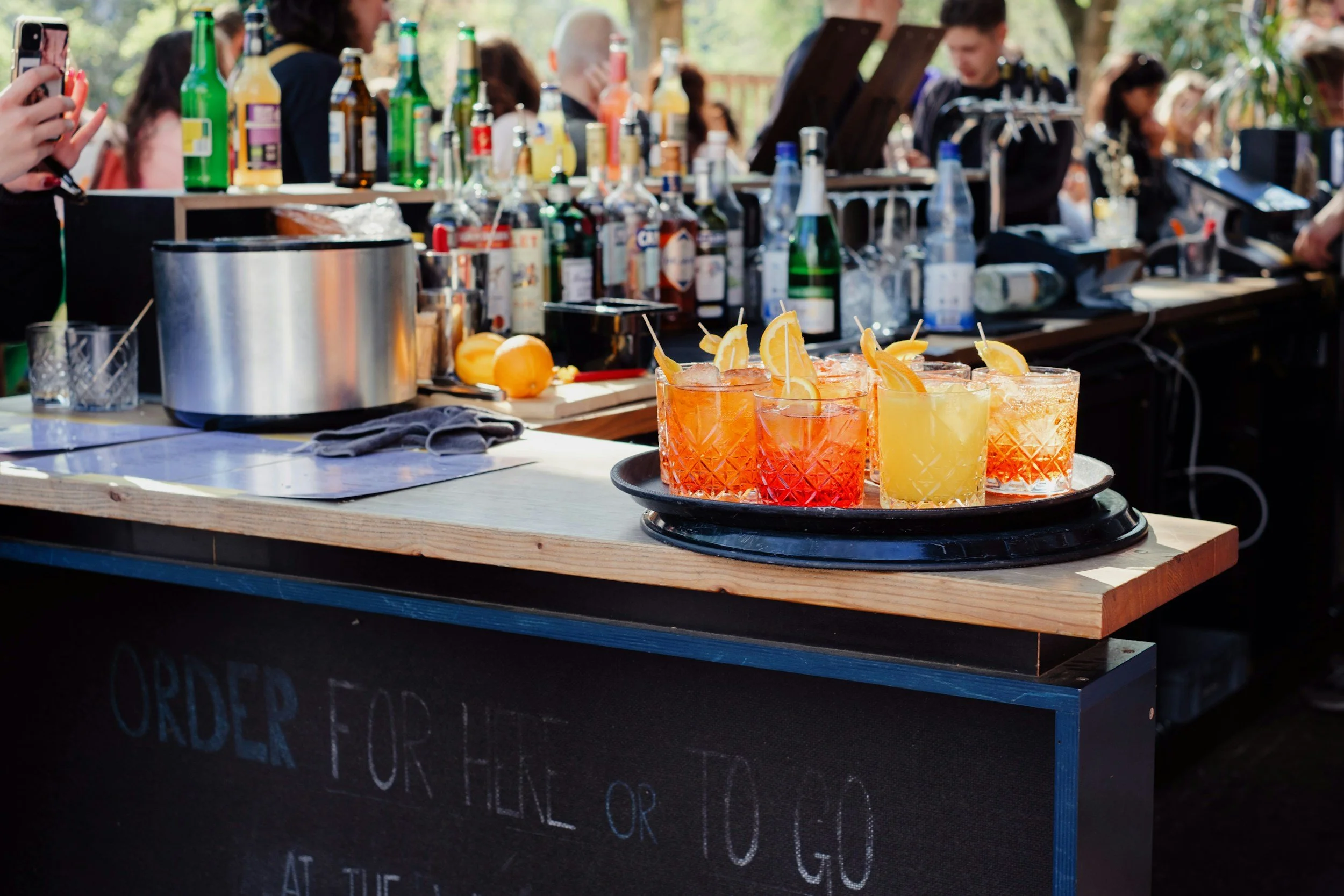A bar setup with various bottles of alcohol, fresh lemons, and colorful cocktails on a black tray, with people in the background socializing outdoors.