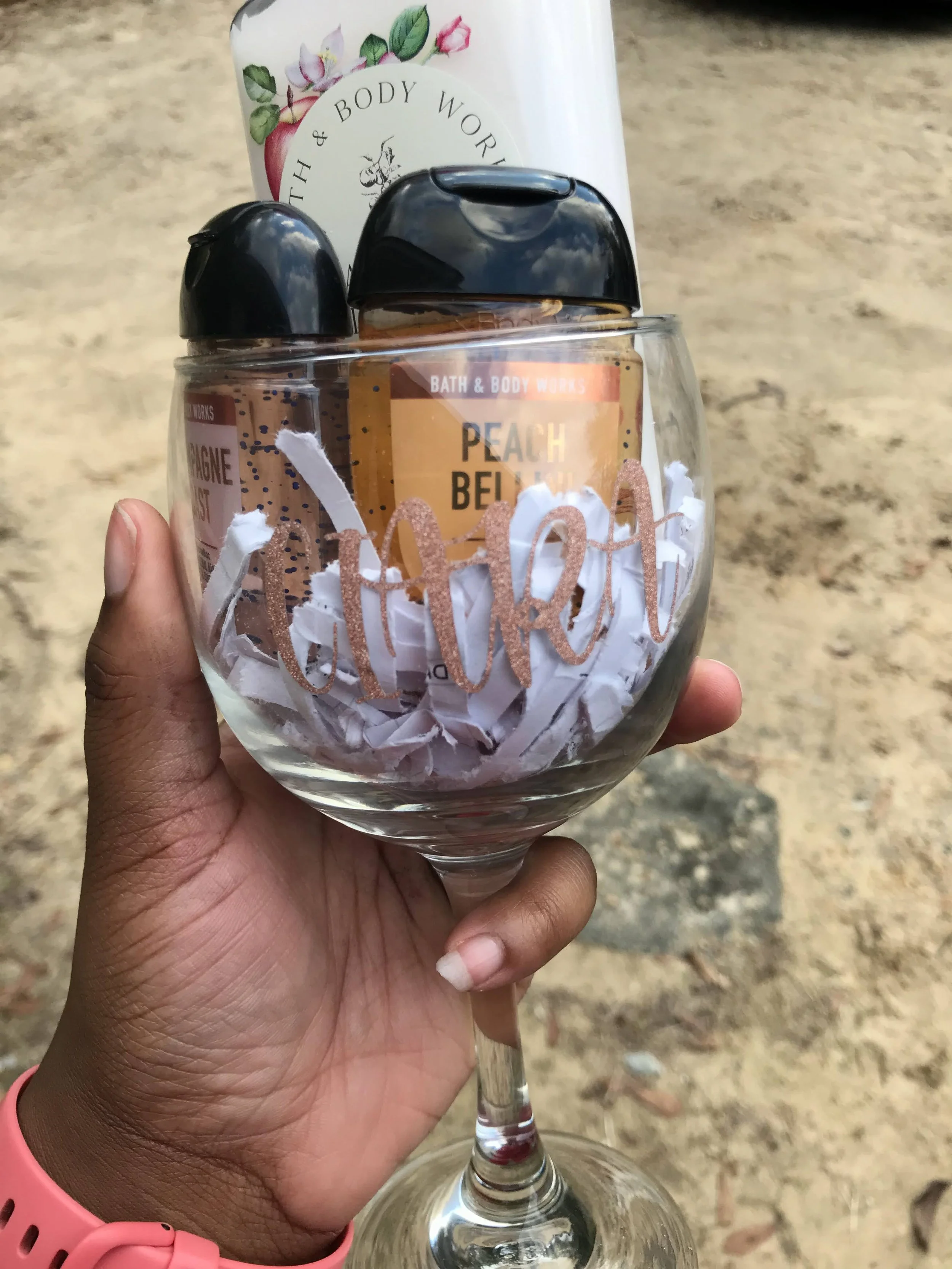 A hand holding a glass of wine with shredded paper and a peach-scented Bath & Body Works hand sanitizer placed on top of it. The background is a blurred outdoor surface.