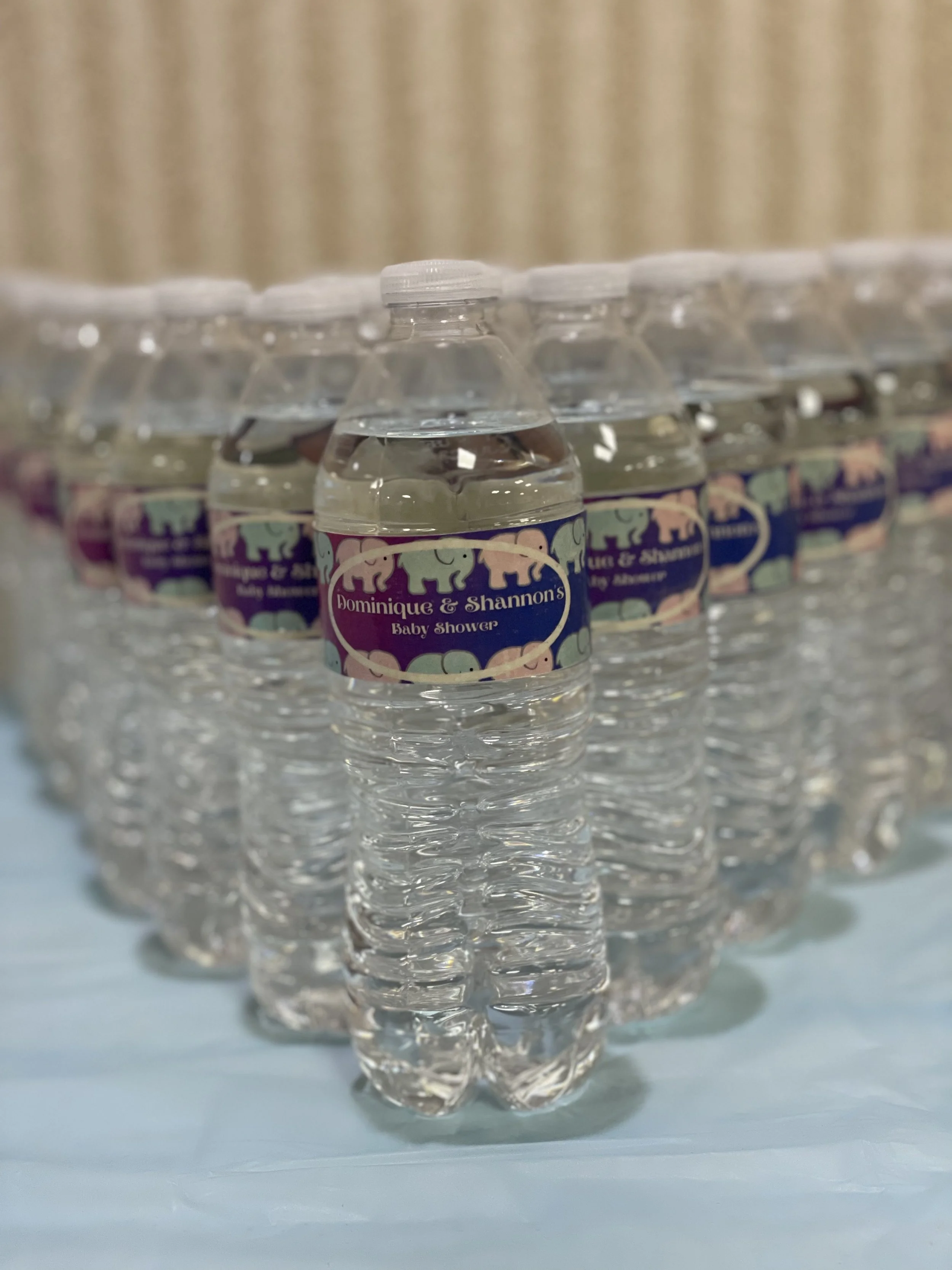 A row of bottled water labeled 'Dominique & Shannon's Baby Shower' with elephant decorations, arranged on a reflective surface.
