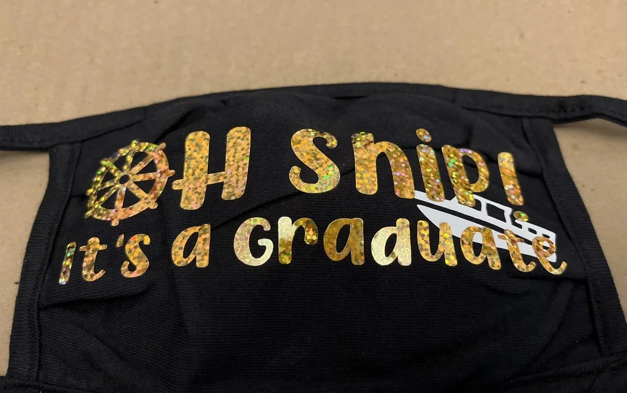 Black face mask with glittery gold text saying 'OH SHIP! it's a graduate' and an emblem of a ship's wheel.