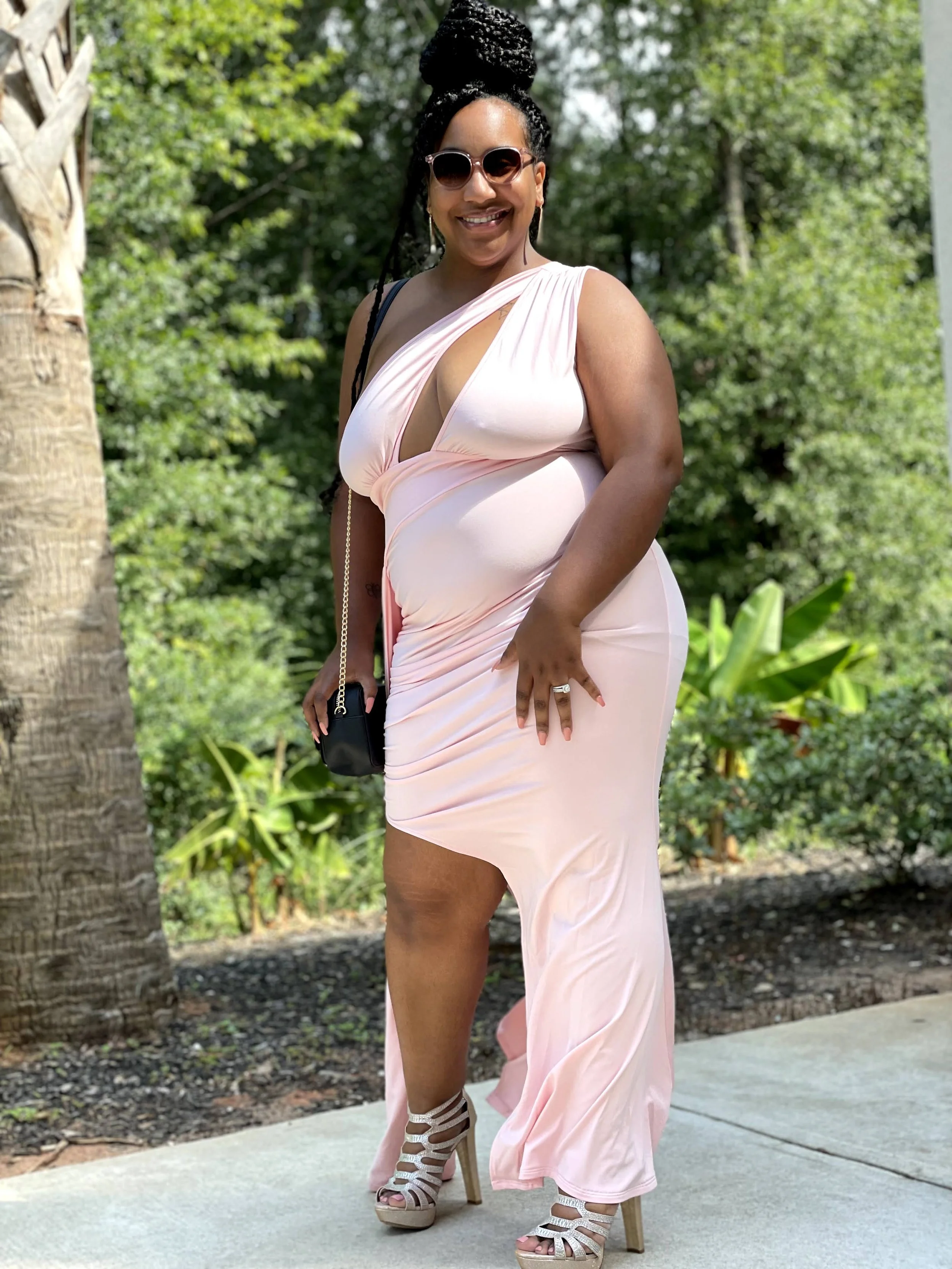 A woman smiling outdoors wearing sunglasses and a light pink dress with a thigh-high slit, standing on a concrete sidewalk with greenery and trees in the background.