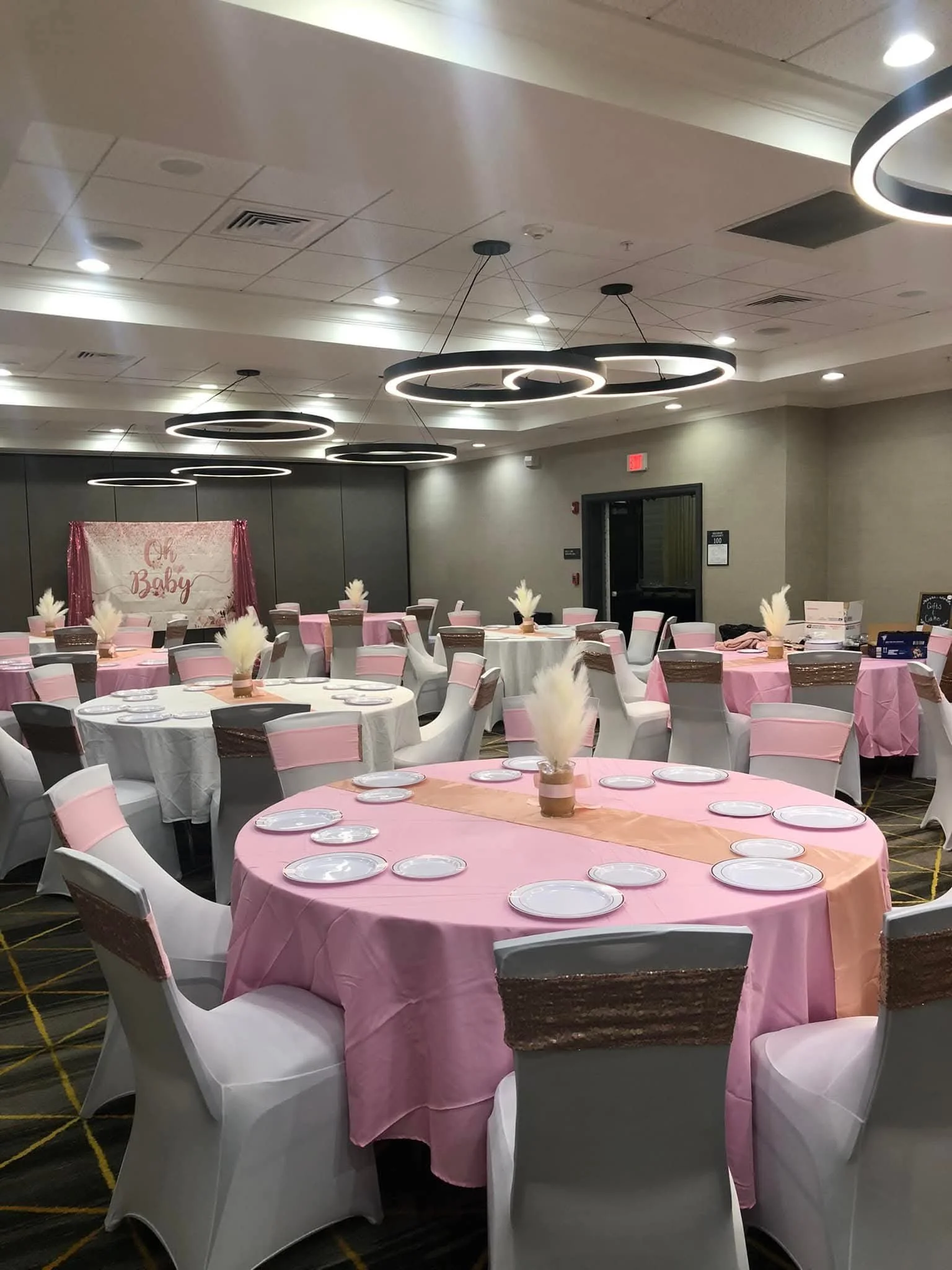 A banquet hall decorated for a baby shower with pink tablecloths, white chairs, and pink and white feather centerpieces. A pink and white banner reads 'Oh Baby' in the background.