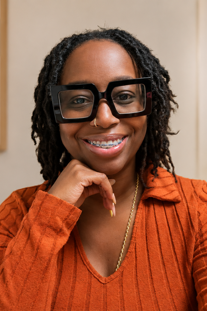 A woman with dark curly hair wearing large black glasses, a nose ring, braces, and an orange textured top, smiling and resting her chin on her hand.