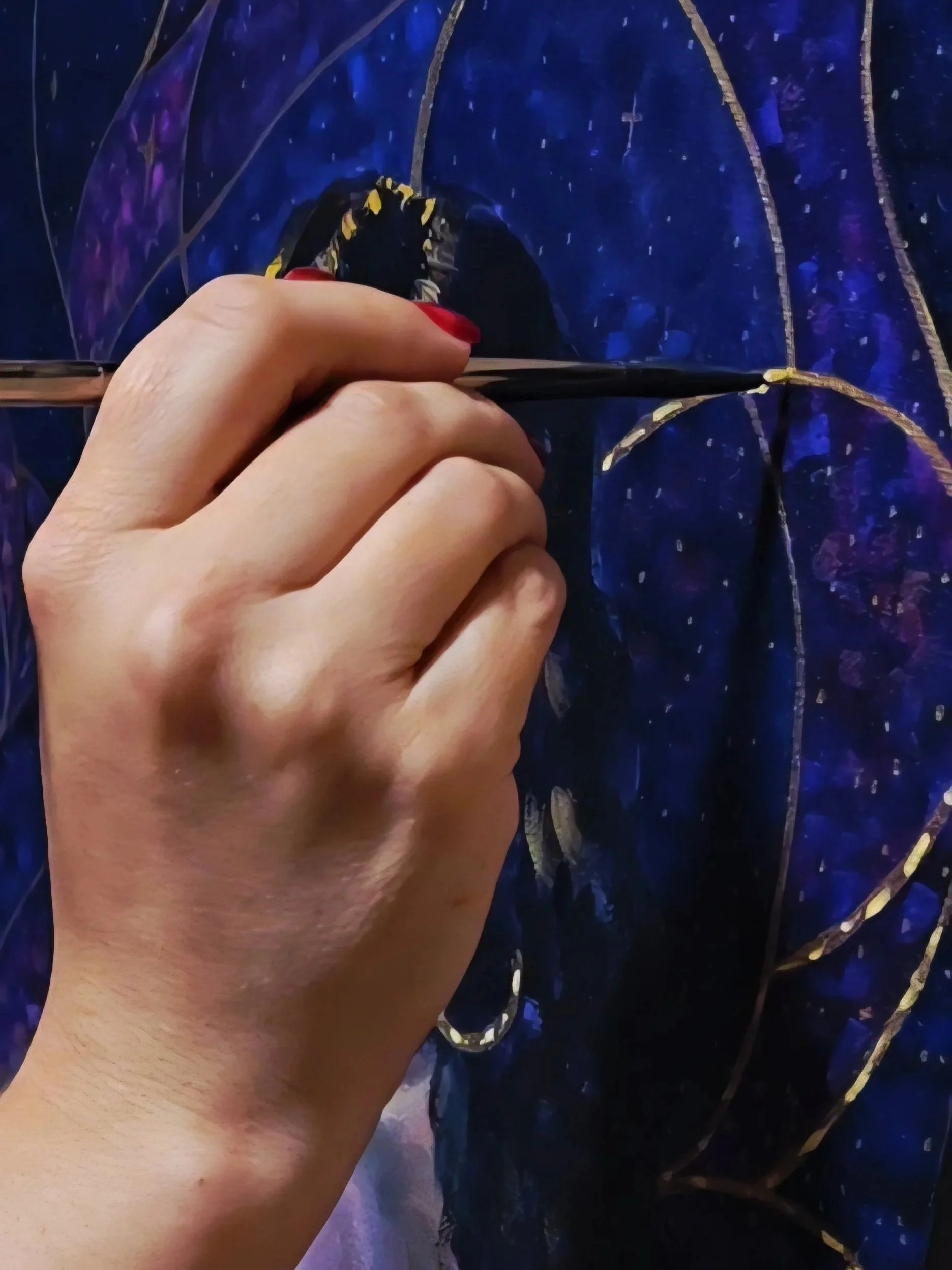 A person's hand with red-painted nails holding a paintbrush and painting details on a dark, vibrant, blue and purple stained glass artwork with gold accents.