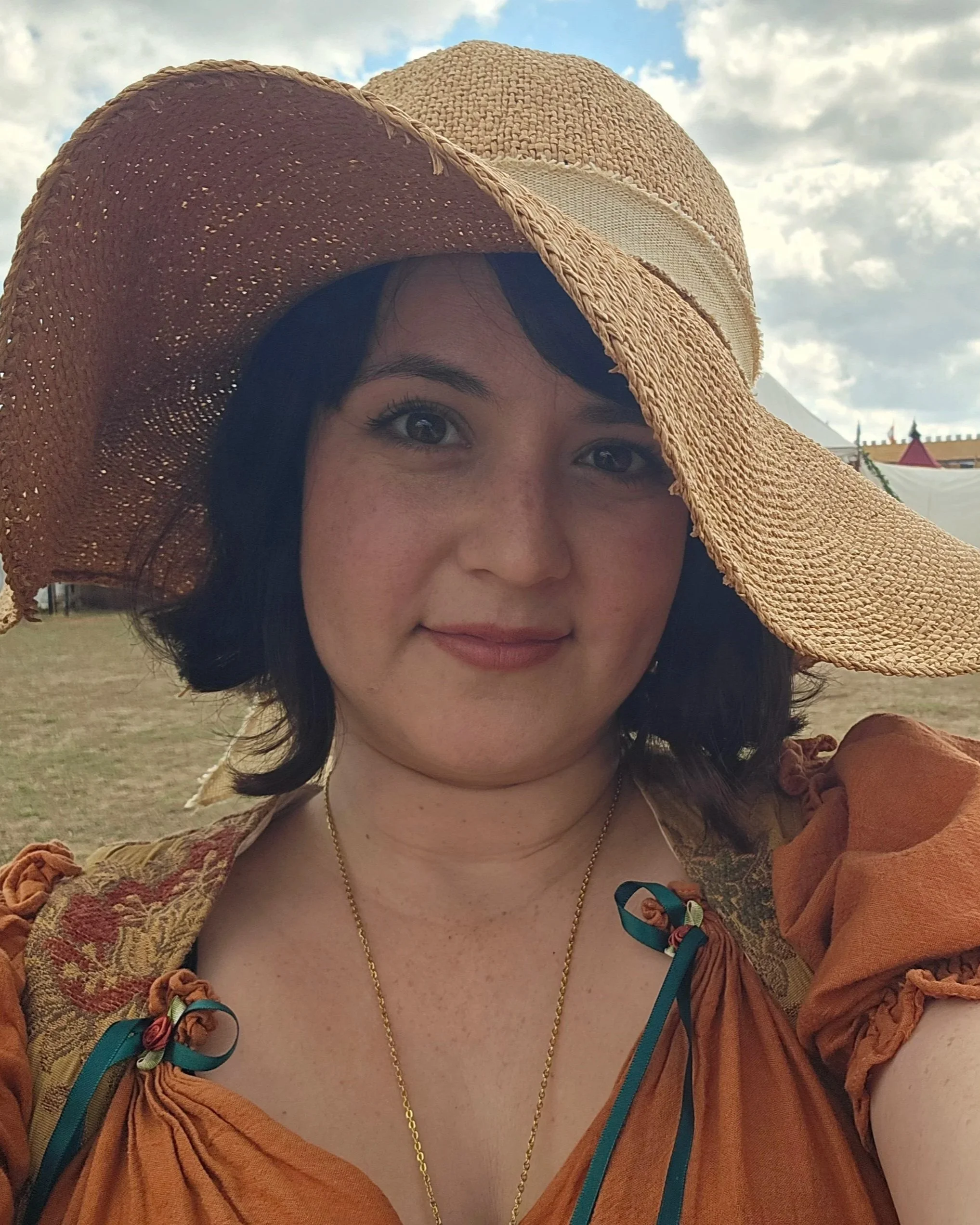 A woman wearing a large woven straw hat with a wide brim, smiling, with dark hair, dark eyes, and gold jewelry, outdoors on a cloudy day.