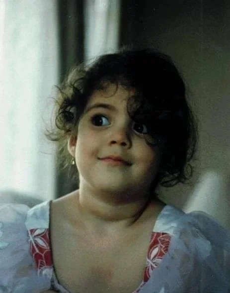 A young girl with curly dark hair and big eyes, wearing a white shirt with red patterned accents on the shoulders, looking slightly to her left indoors near a window with white curtains.