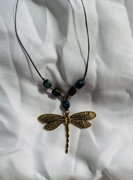 Gold dragonfly pendant necklace with black and teal beads on a black cord, displayed on white fabric.
