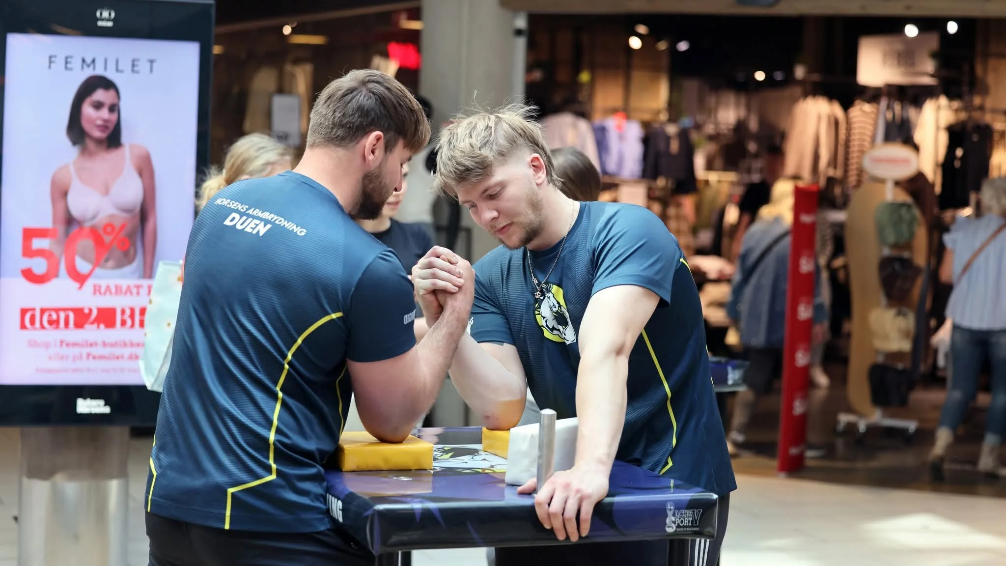 To unge mænd deltager i en armstrækkonkurrence i en butik, begge iført sportstrøjer med logoer. Baggrund med butiksskyggelokaler og reklameskilte.