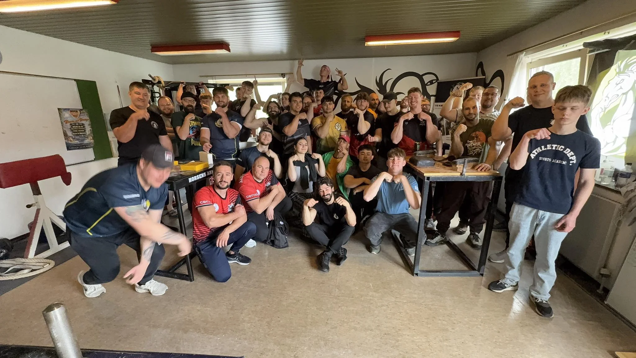 En stor gruppe mennesker, der poser for et gruppbillede indendørs. Nogle er muskuløse og viser armbracelet, og de fleste smiler. Der er både voksne og børn, og nogle står ved borde, mens andre står bagved.