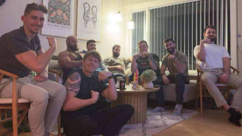 Syv mænd sidder i en stue, nogle viser deres muskler, og der er cola på bordet, samt blomsterdekoration.