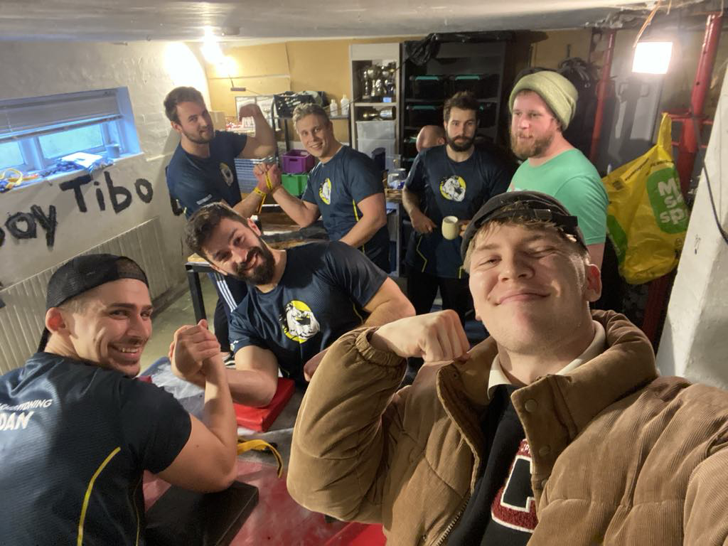 Syv mænd, nogle i sportstrøjer, samles indendørs med værktøjskasser og dagligdags ting i baggrunden. De ser glade ud og laver en armstrækning eller flexer musklerne, mens de tager et gruppe selfie.