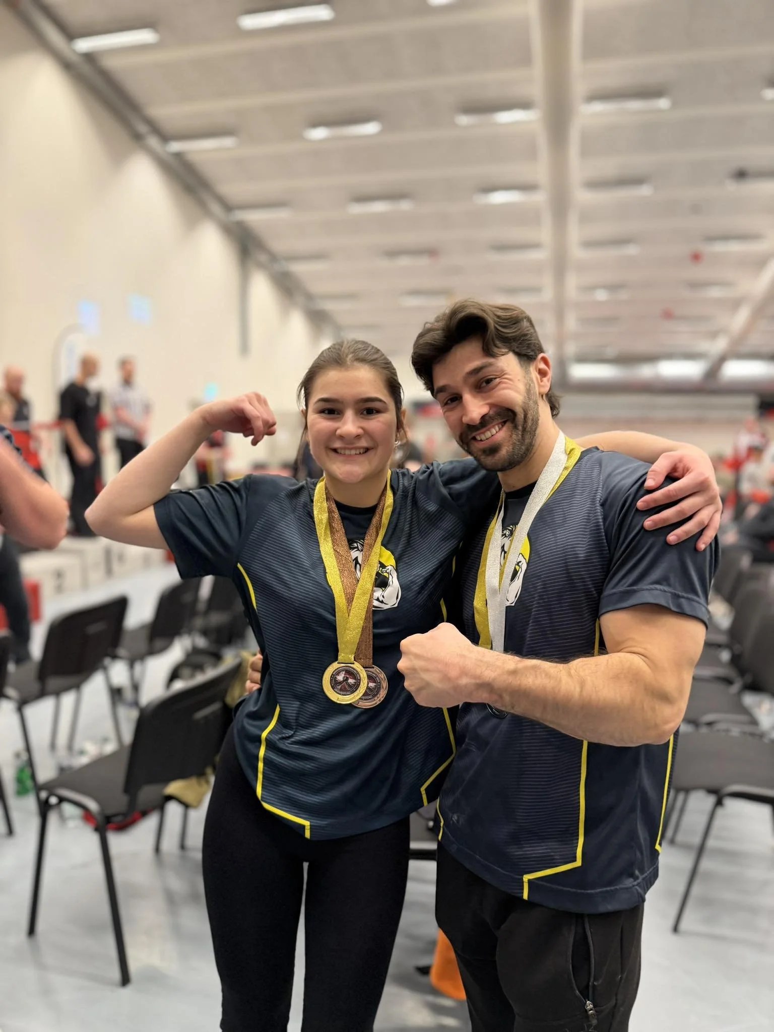 To personer i sporttøj smiler og viser medaljer, indendørs med andre mennesker og sportsudstyr i baggrunden.